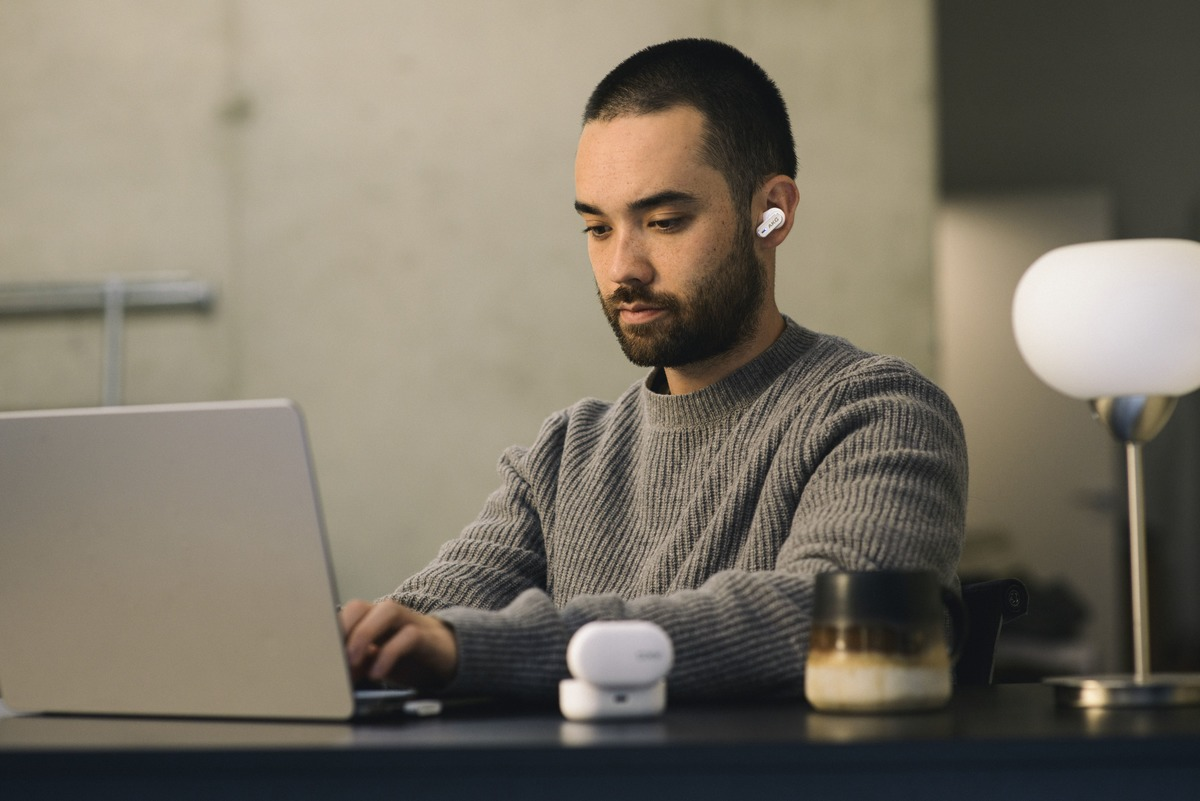Mann mit Ohrhörern arbeitet am Laptop, weiße Ohrhörerbox und Lampe auf Schreibtisch.