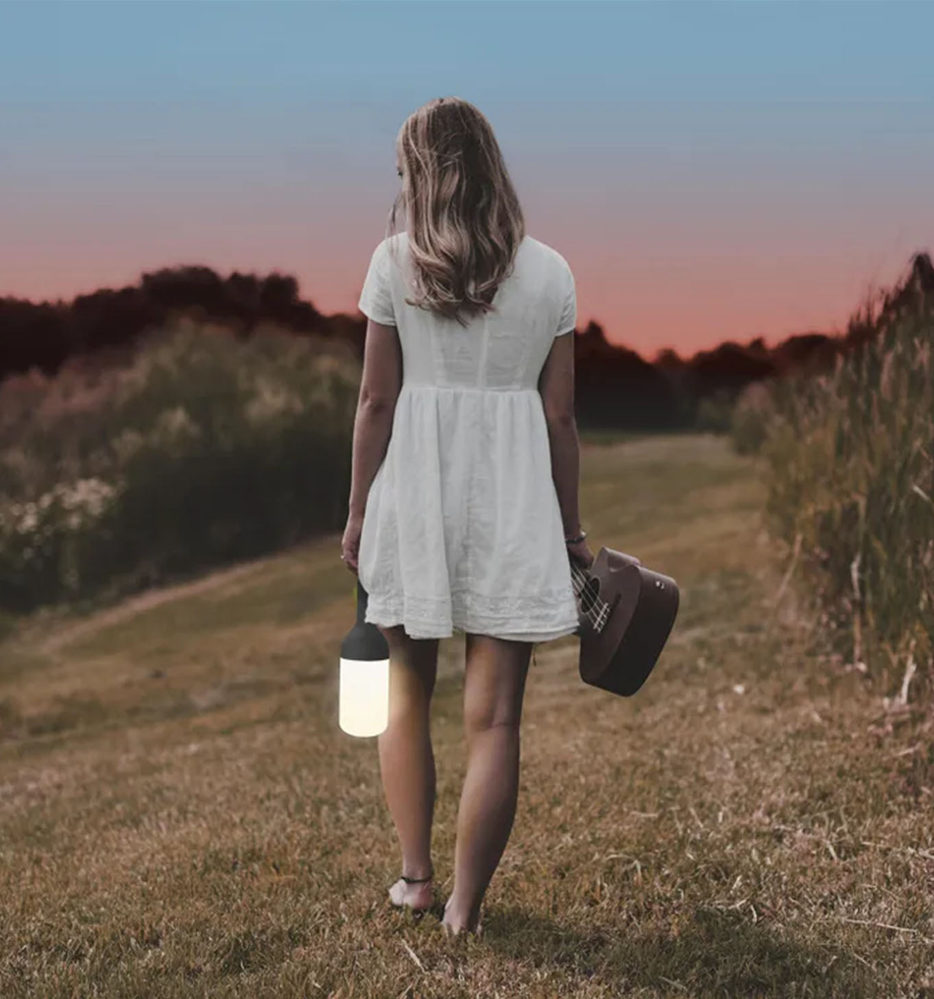 Frau in weißem Kleid geht mit Laterne und Ukulele bei Sonnenuntergang.