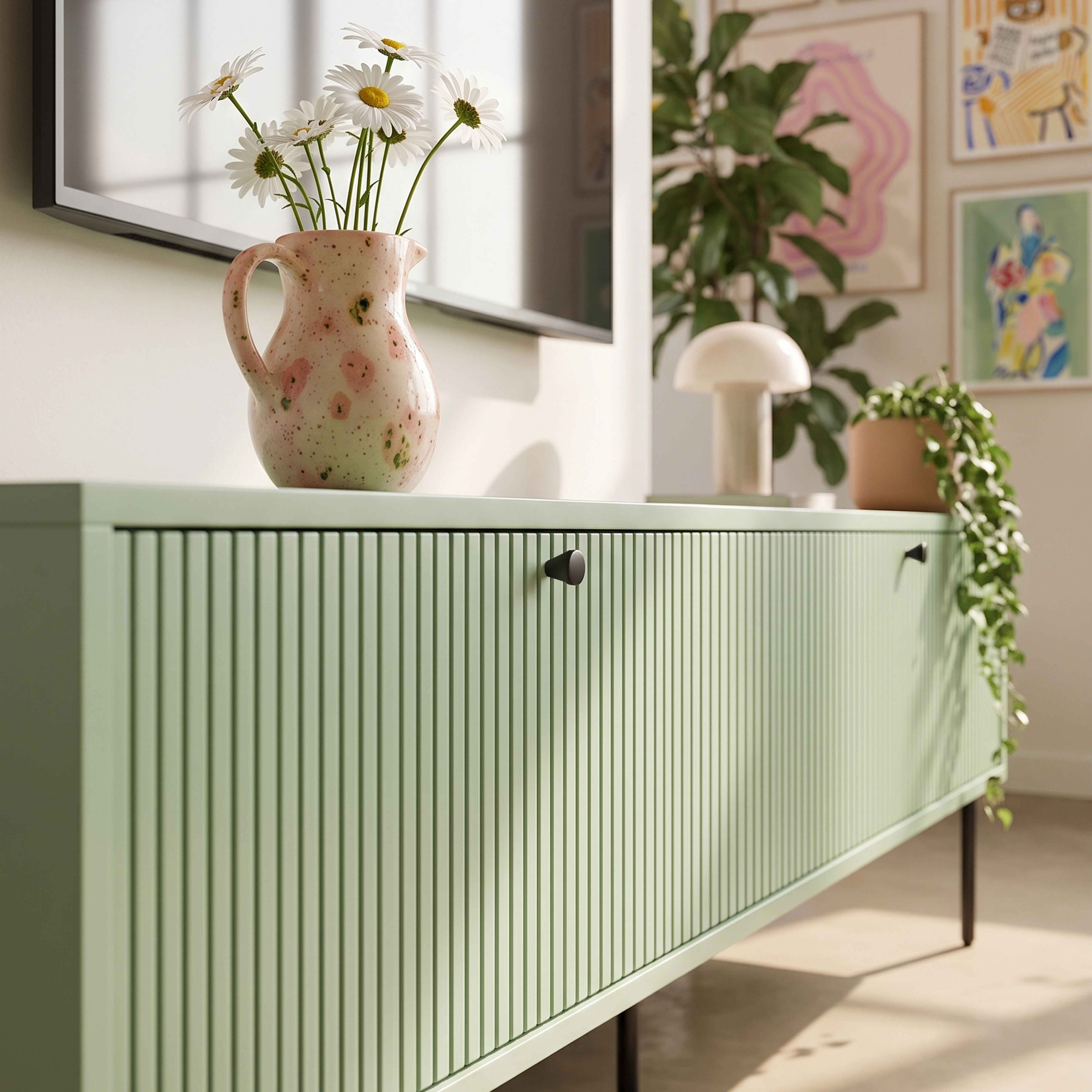 Ein hellgrünes Sideboard mit Rillenfront und schwarzen Griffen, darauf eine Vase mit Gänseblümchen.