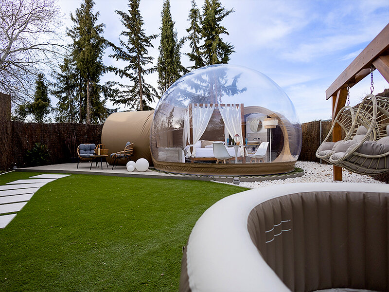 Una carpa burbuja inflable con cama y mesa, junto a asientos y columpios colgantes.
