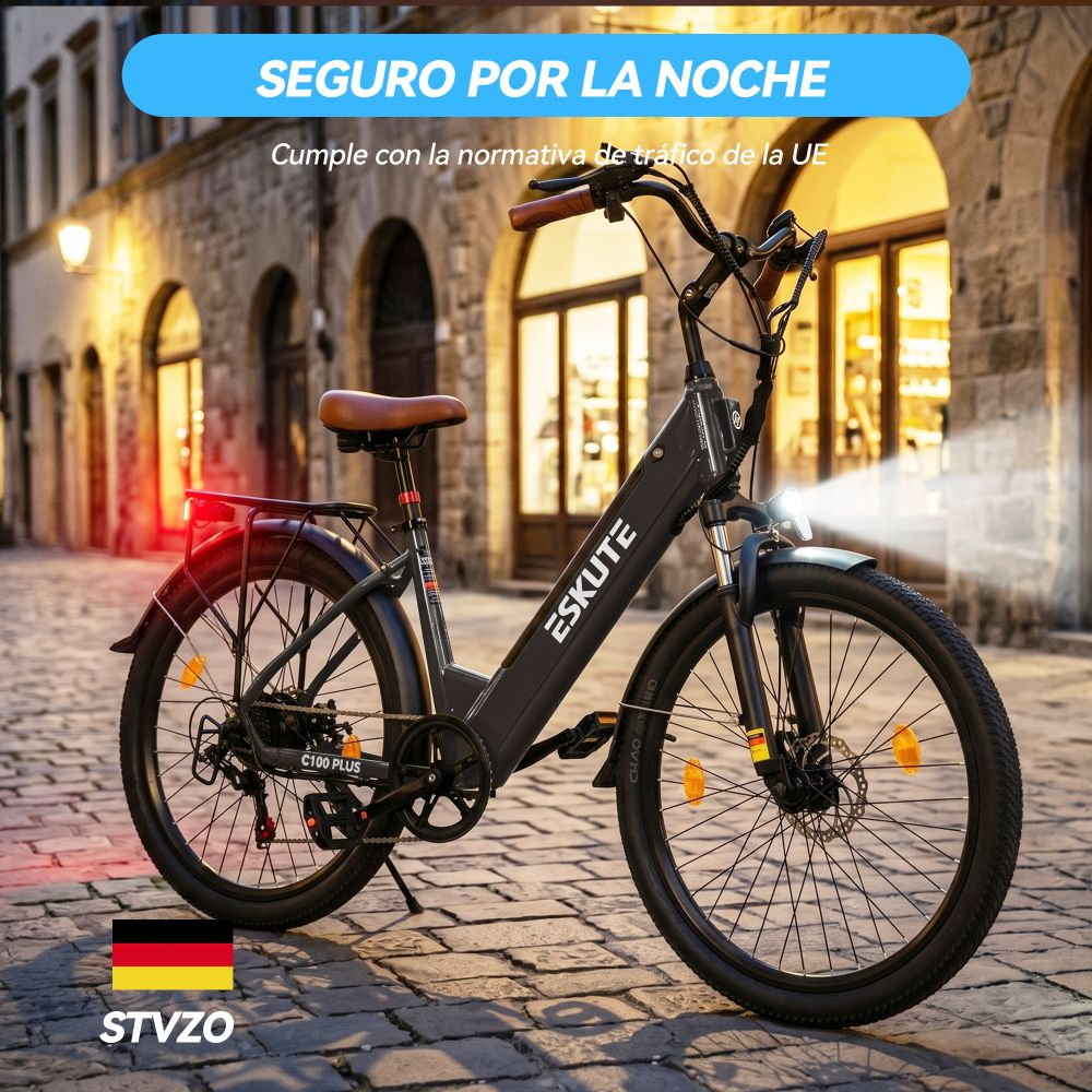 Una bicicleta eléctrica oscura está aparcada sobre adoquines frente a edificios iluminados. Tiene sillín y manillar marrones.
