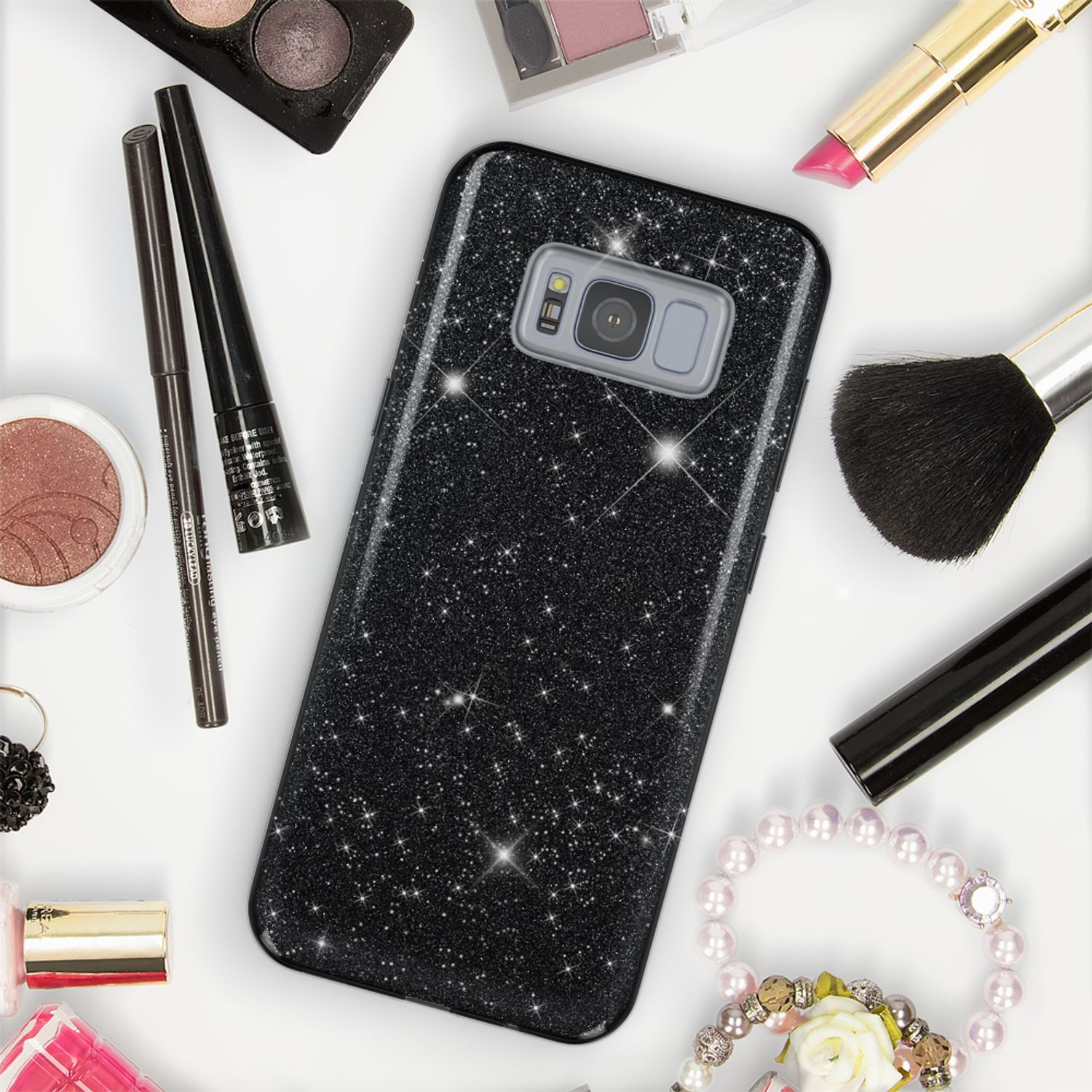 Schwarze glitzernde Handyhülle umgeben von Kosmetika wie Lippenstift, Lidschatten, Pinsel, Perlenarmband.