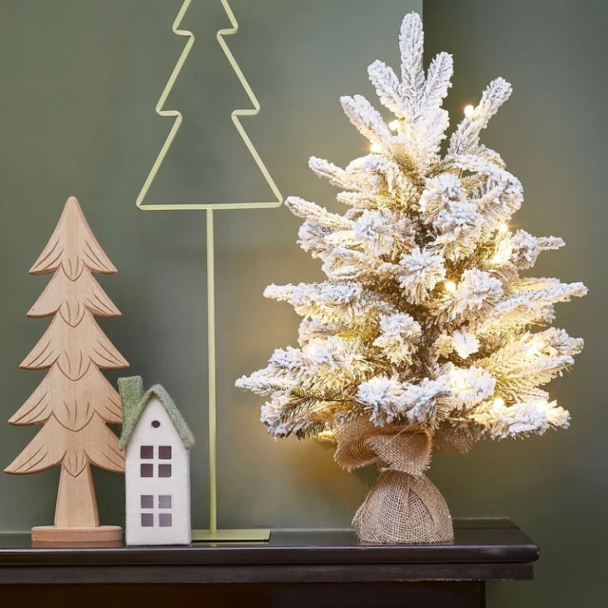 A lit snowy Christmas tree with a jute base, next to a wooden figure and house.
