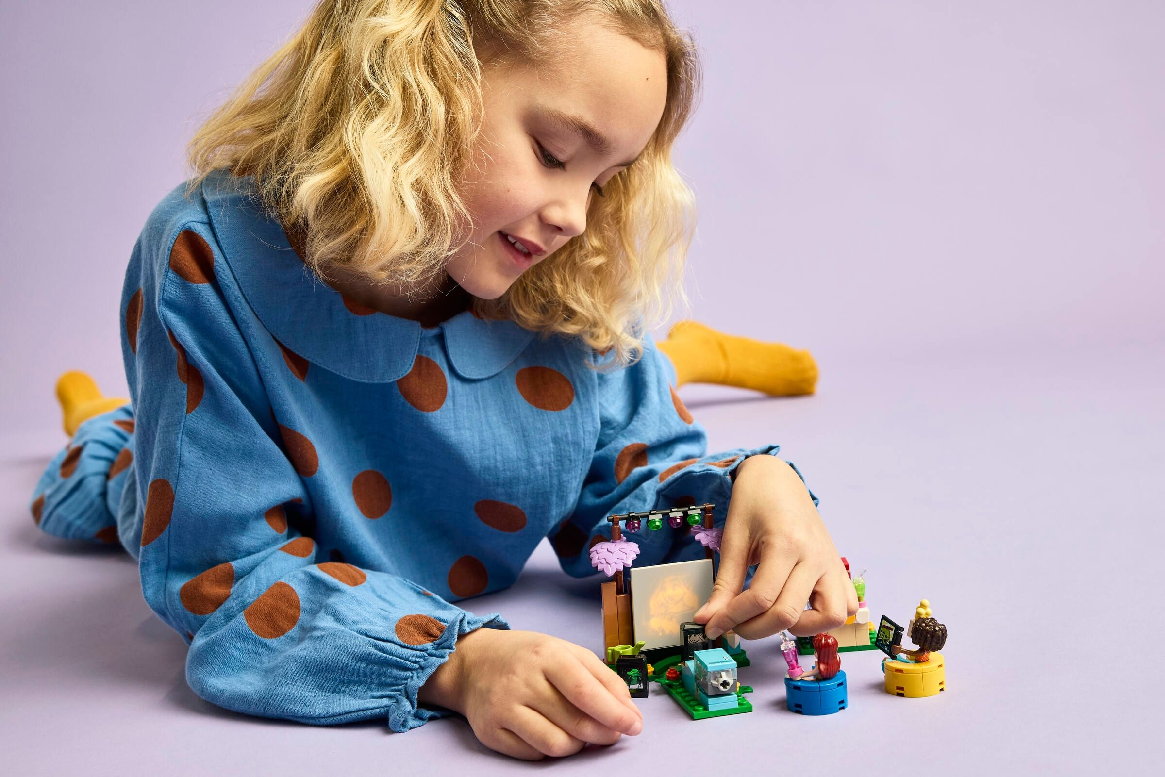 Ein Mädchen in einem blauen gepunkteten Kleid spielt mit einem Lego-Set auf lila Hintergrund.