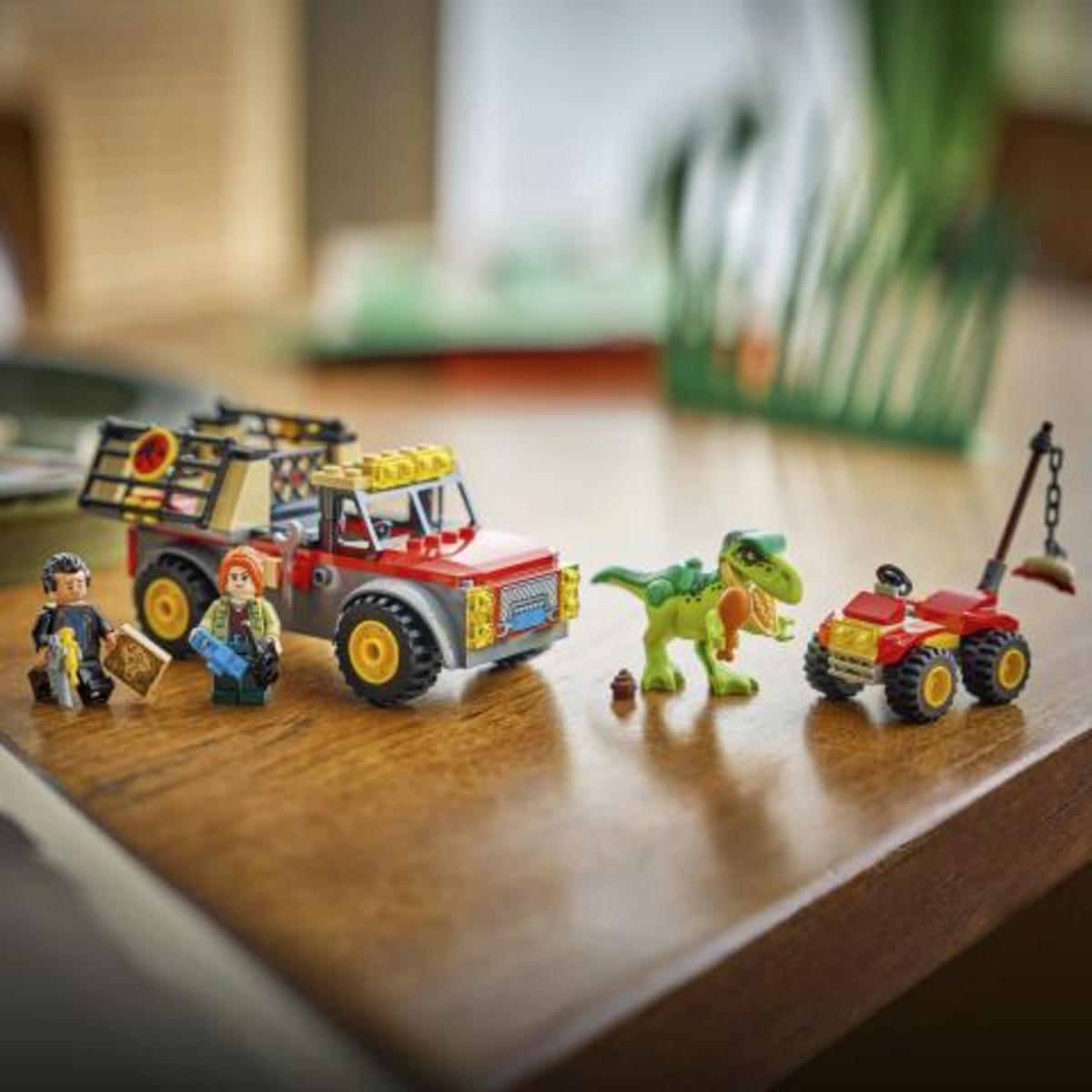 Due figure Lego accanto a un camion rosso e grigio con gabbia. Un dinosauro verde e un veicolo più piccolo.