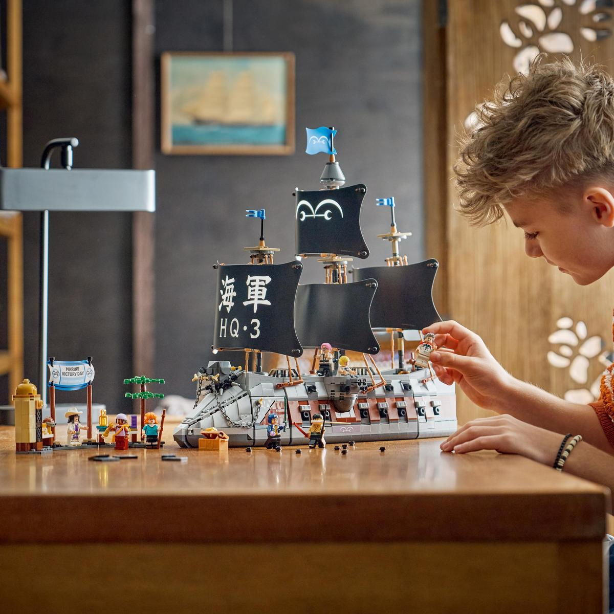 Niño construye un barco Lego con velas negras y figuras sobre mesa de madera.