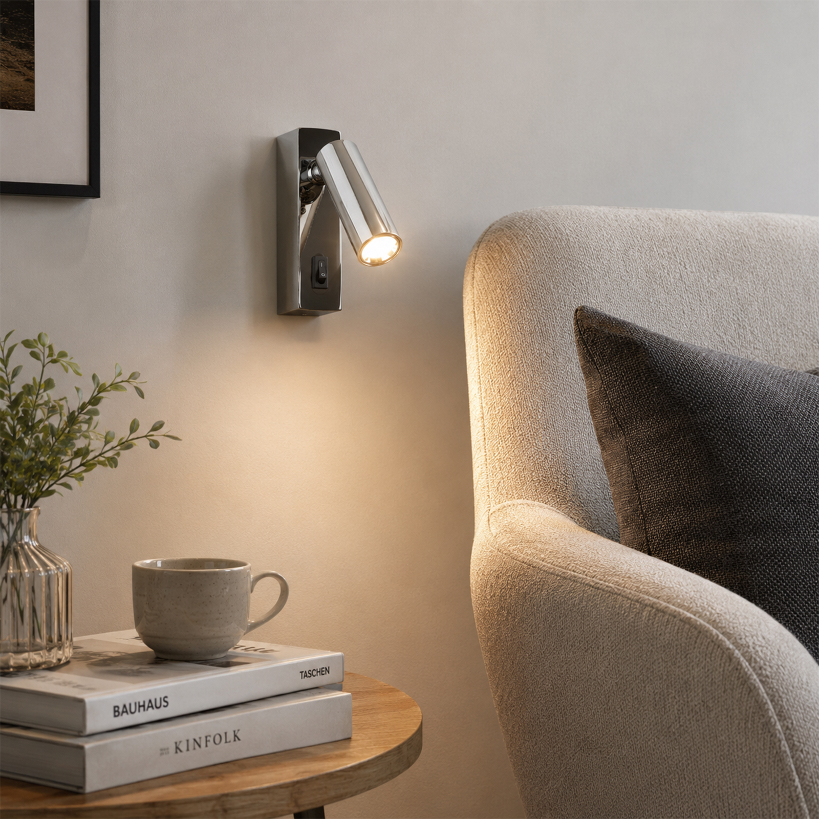 Un aplique plateado ilumina un sillón con cojines. Al lado, una mesa con libros y jarrón.