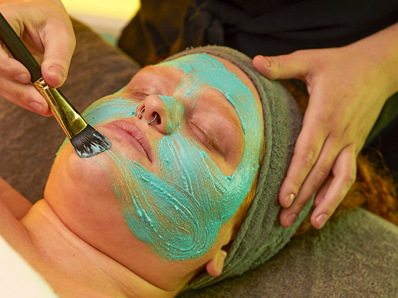 Una persona aplica una mascarilla turquesa con un pincel en la cara de otra persona.