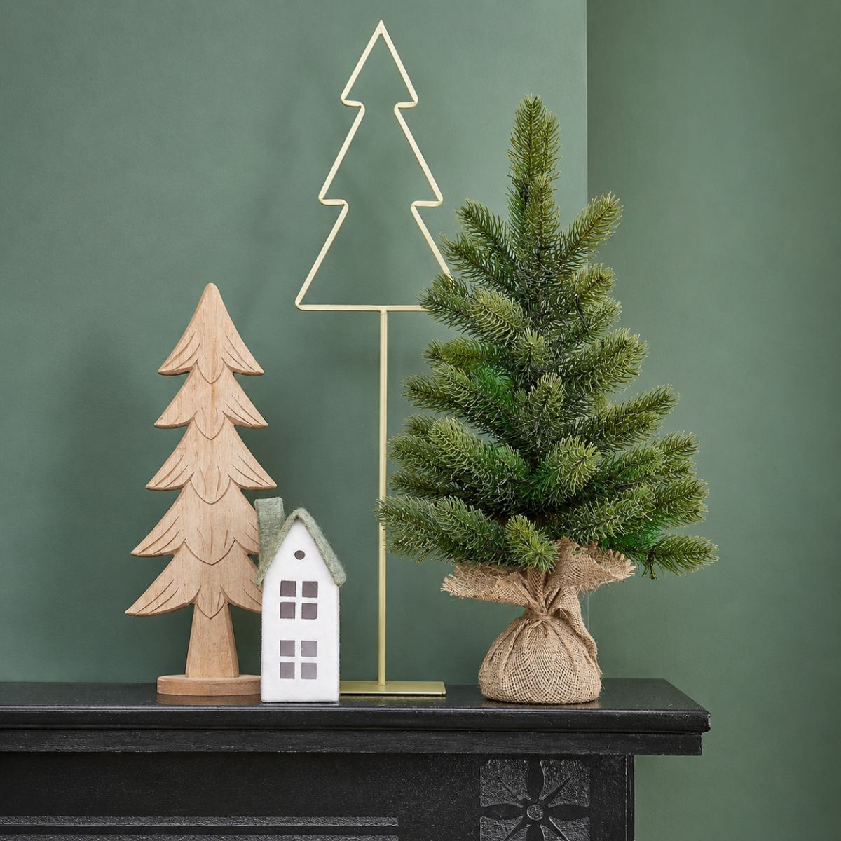 Christmas display on black mantelpiece: wooden tree, white house, gold tree outline, green tree in burlap.