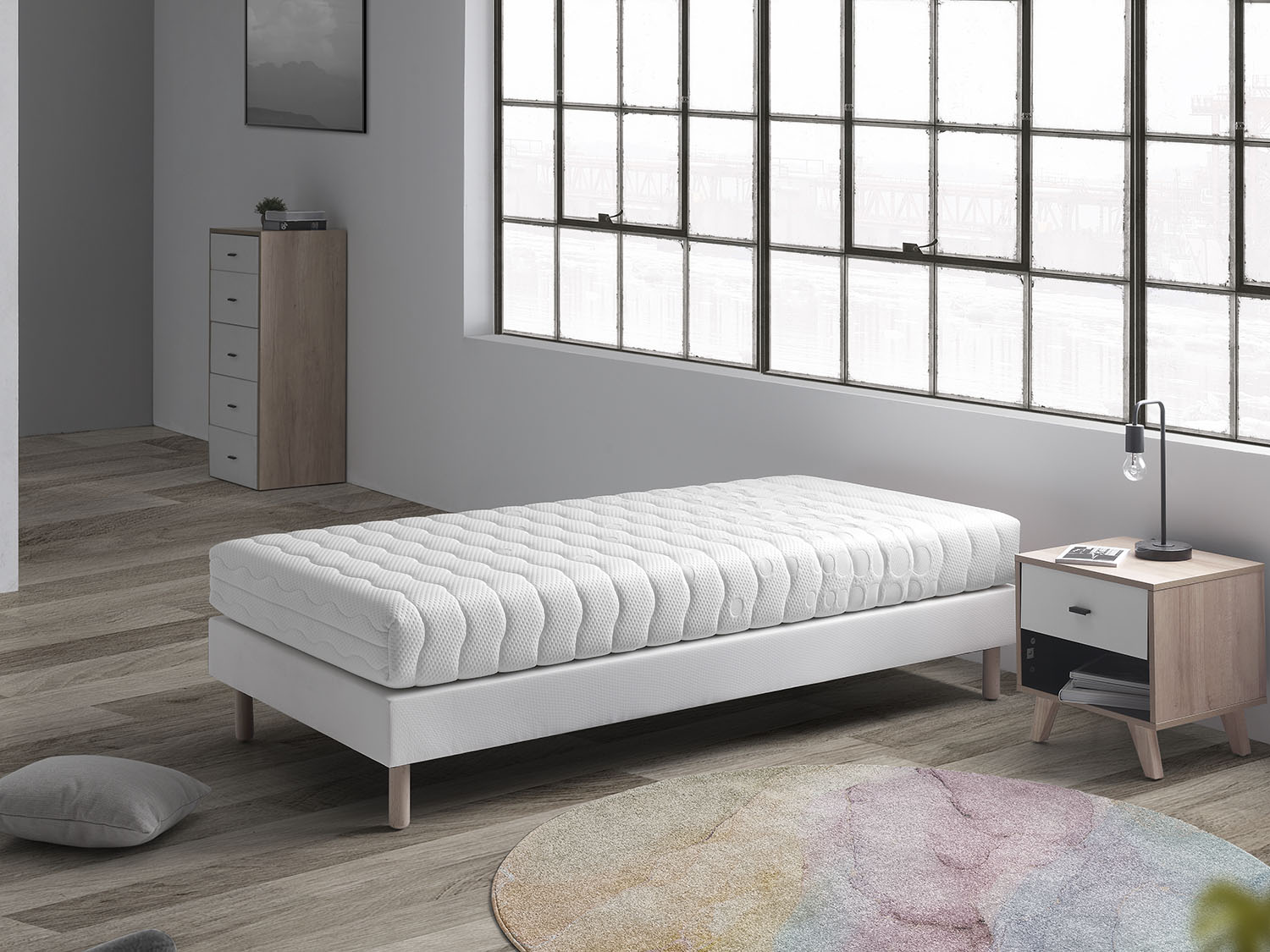 Una cama blanca con colchón se alza en una habitación con suelo de madera y ventana grande.