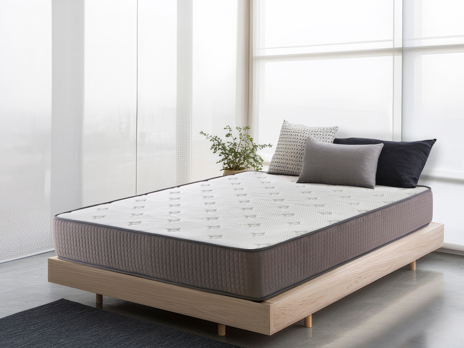 Una cama con colchón blanco, tres almohadas y una planta.