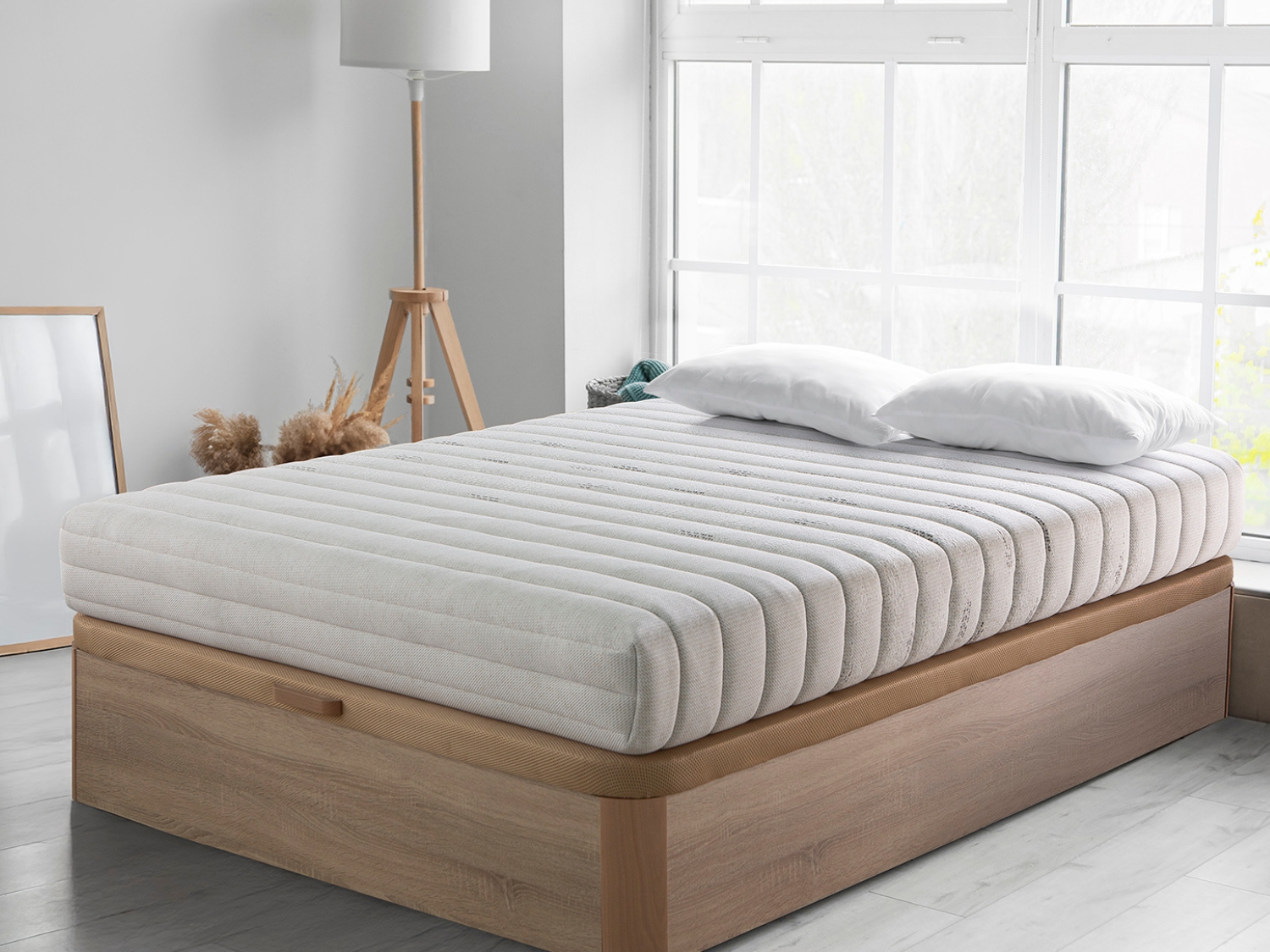 Una cama con colchón blanco, dos almohadas, marco de madera y ventana al fondo.