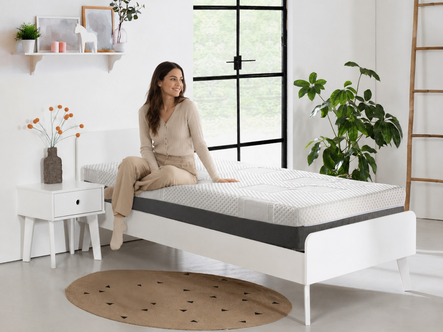 Mujer sentada en cama blanca con colchón, mesita de noche, alfombra y ventana.
