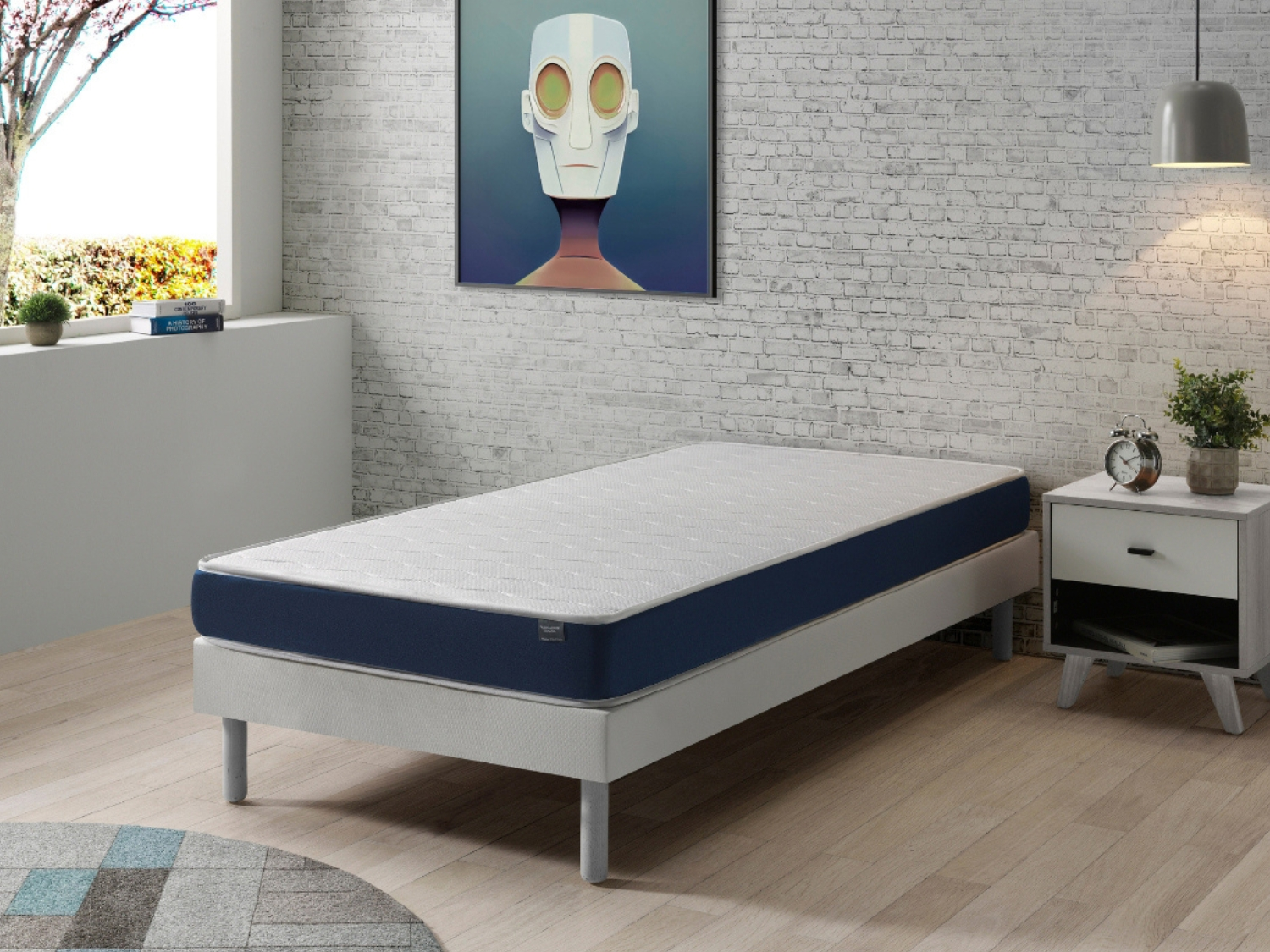 Una cama con borde azul sobre suelo de madera contra pared de ladrillos.