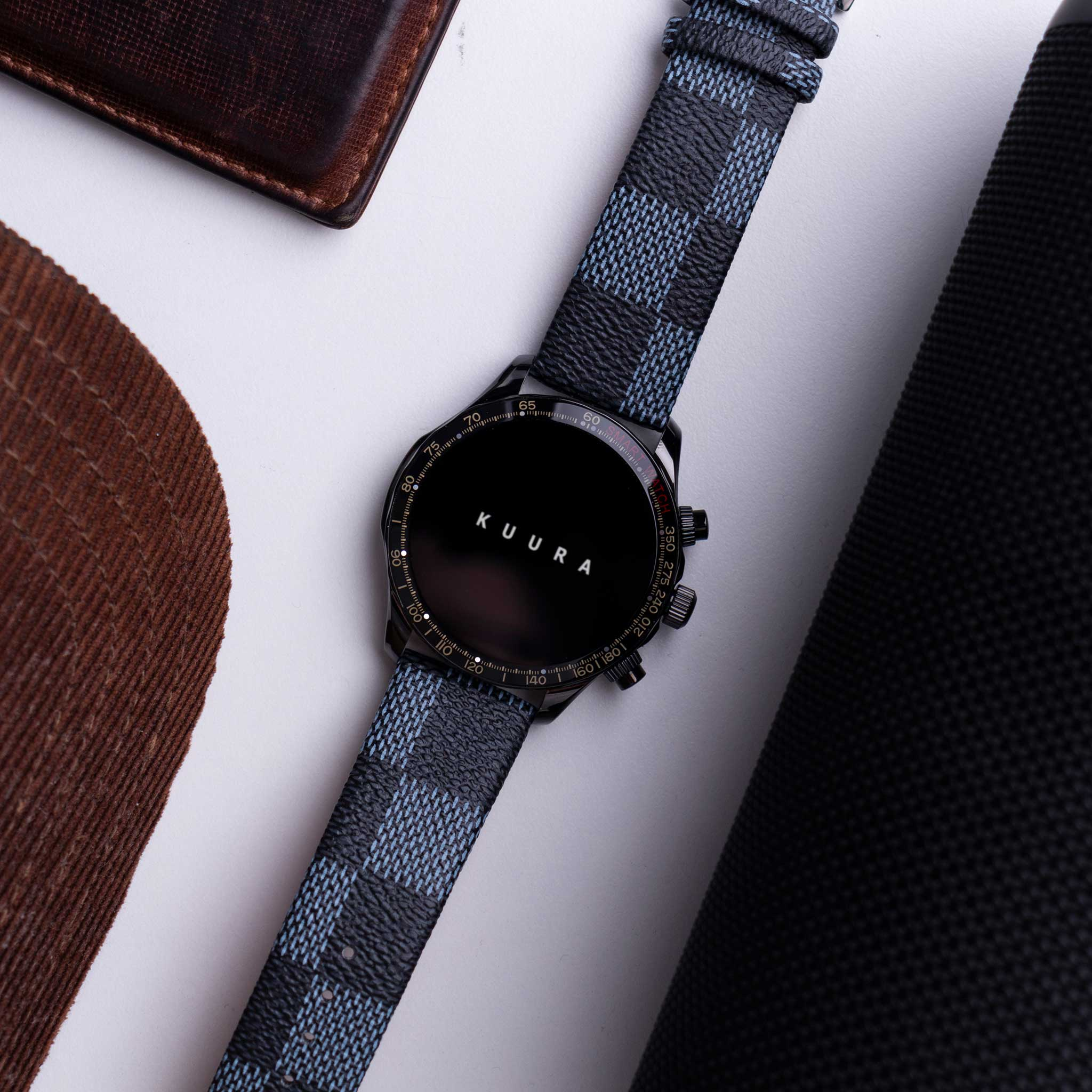 Czarny smartwatch w niebieską kratę leży obok brązowych skórzanych przedmiotów.