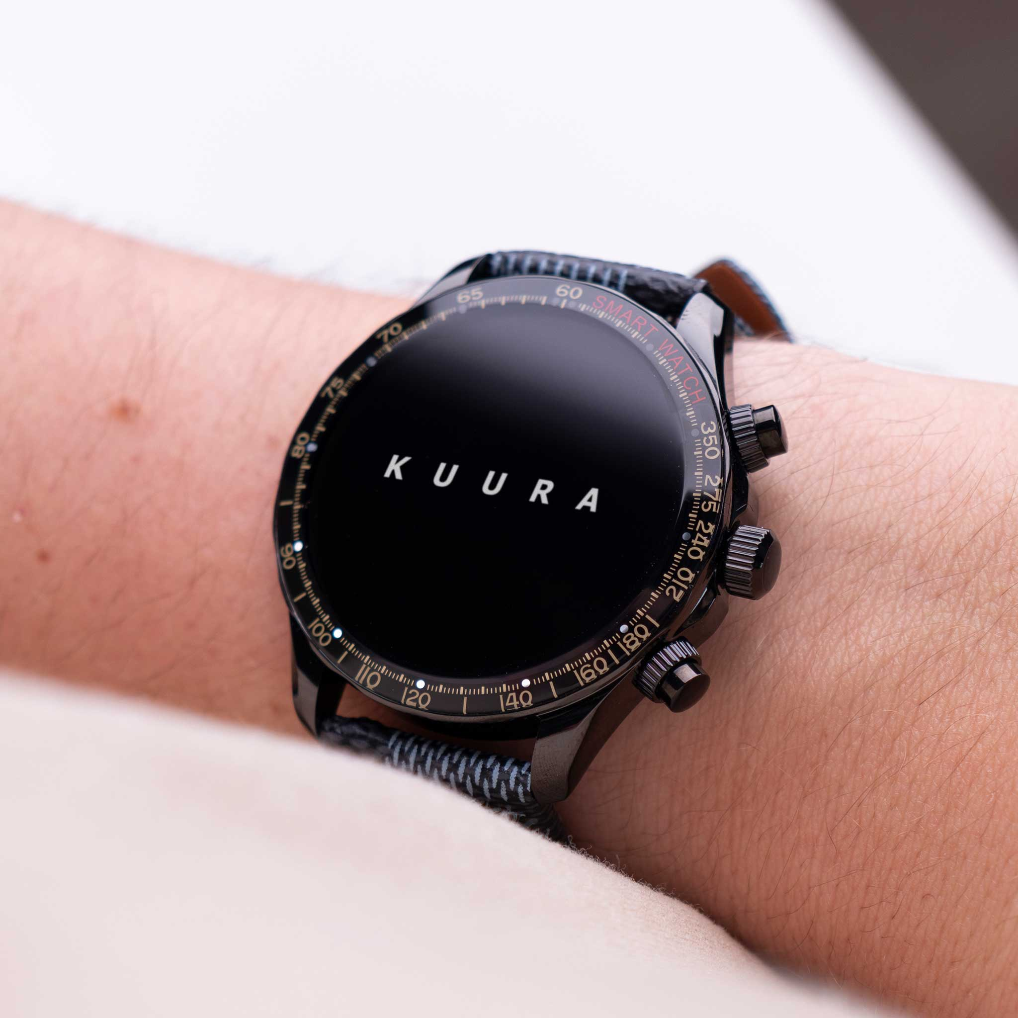 Czarny smartwatch ze srebrnymi oznaczeniami i napisem "KUURA" jest noszony na ramieniu.
