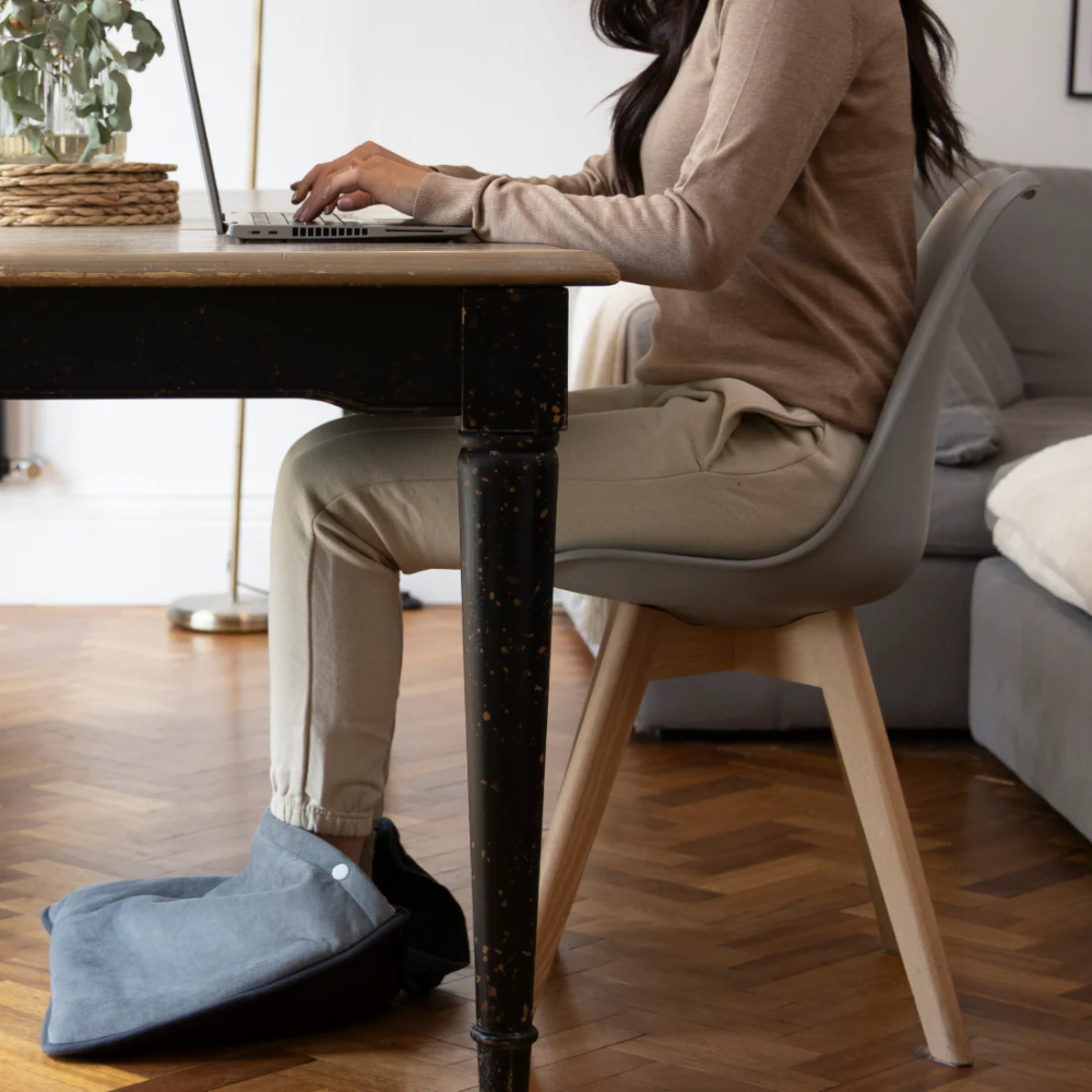 Une femme assise à une table tape sur un ordinateur portable. Ses pieds sont dans un repose-pieds gris.