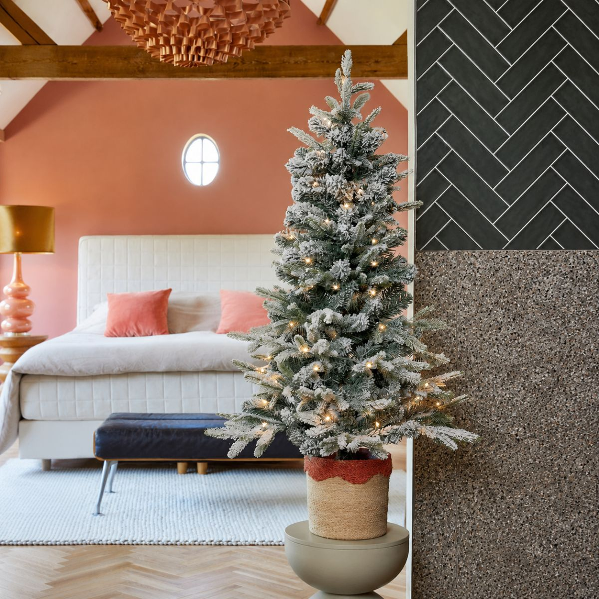 A flocked Christmas tree with lights sits in a basket, in front of a bed.