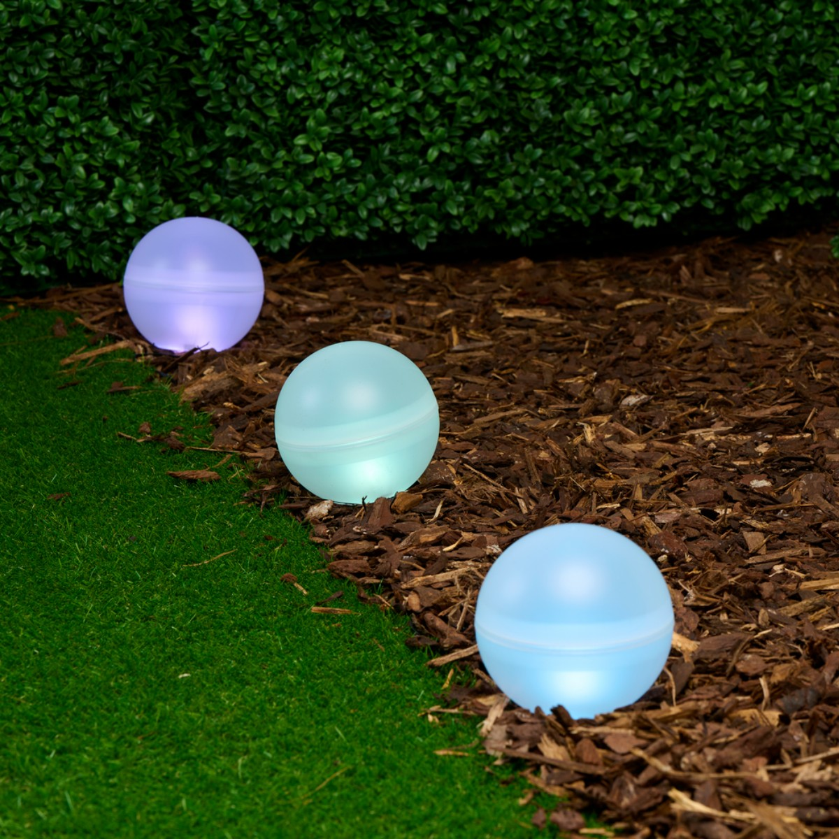 Drie verlichte bollen in paars, cyaan en blauw liggen op mulch naast gras.