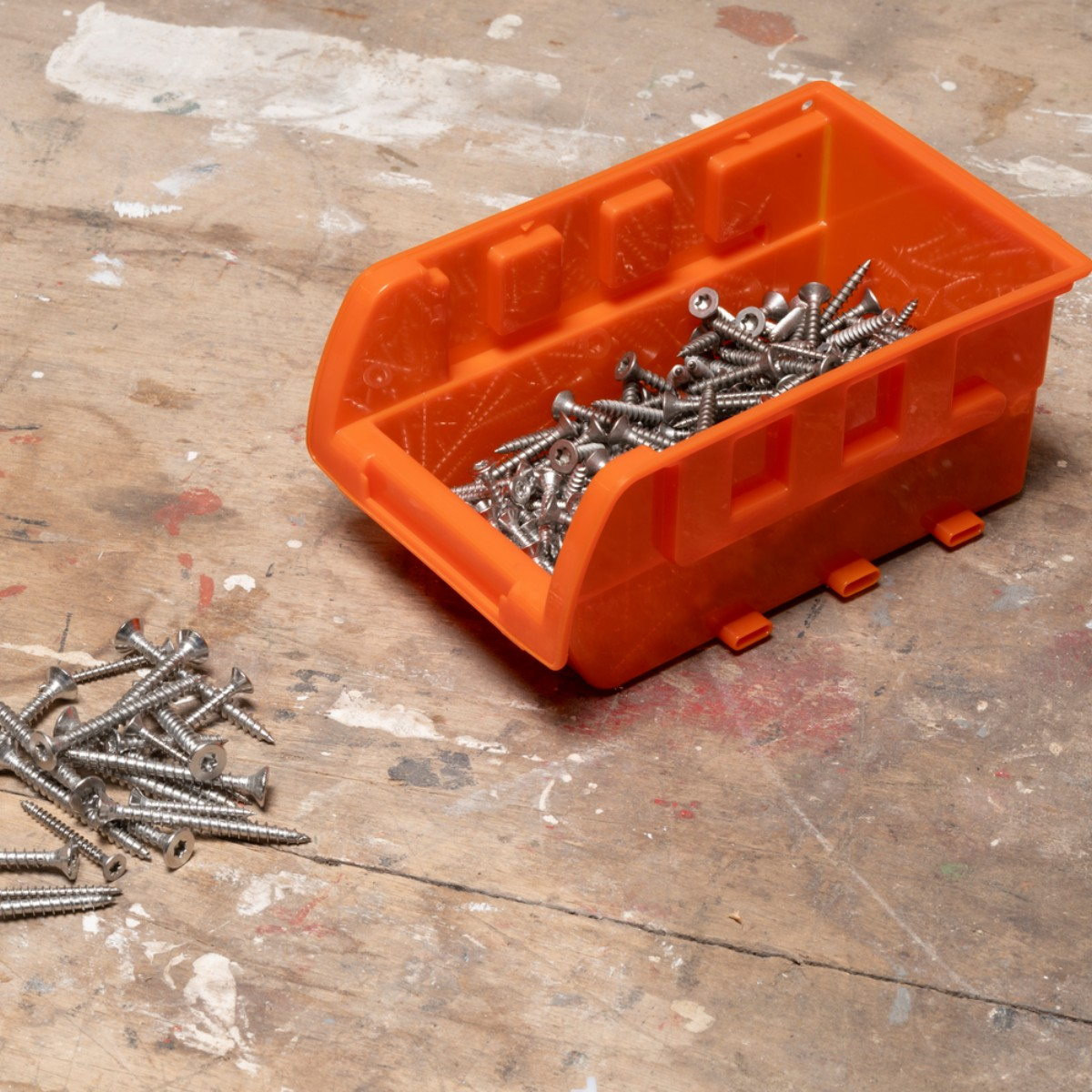 Ein orangefarbener Behälter mit Schrauben und ein Haufen Schrauben daneben auf einem Holztisch.