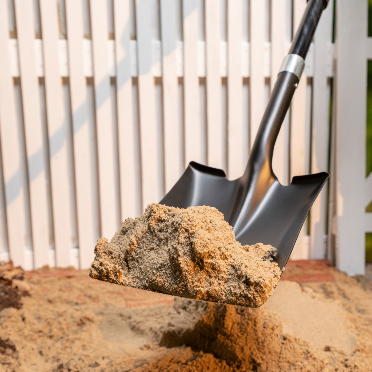 A black shovel lifts sand in front of a white fence.