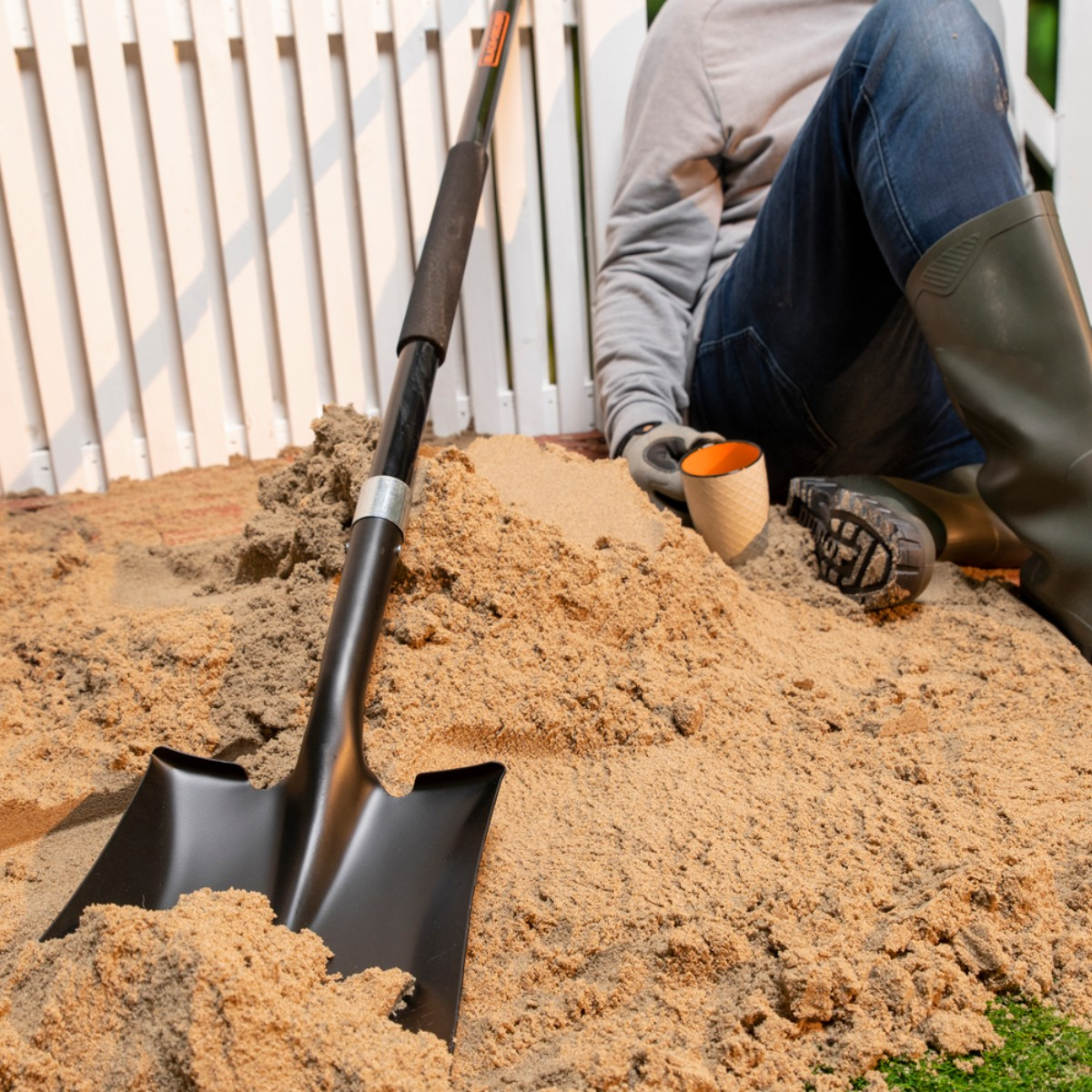 Shovel in sand, person sitting nearby with boots and cup.