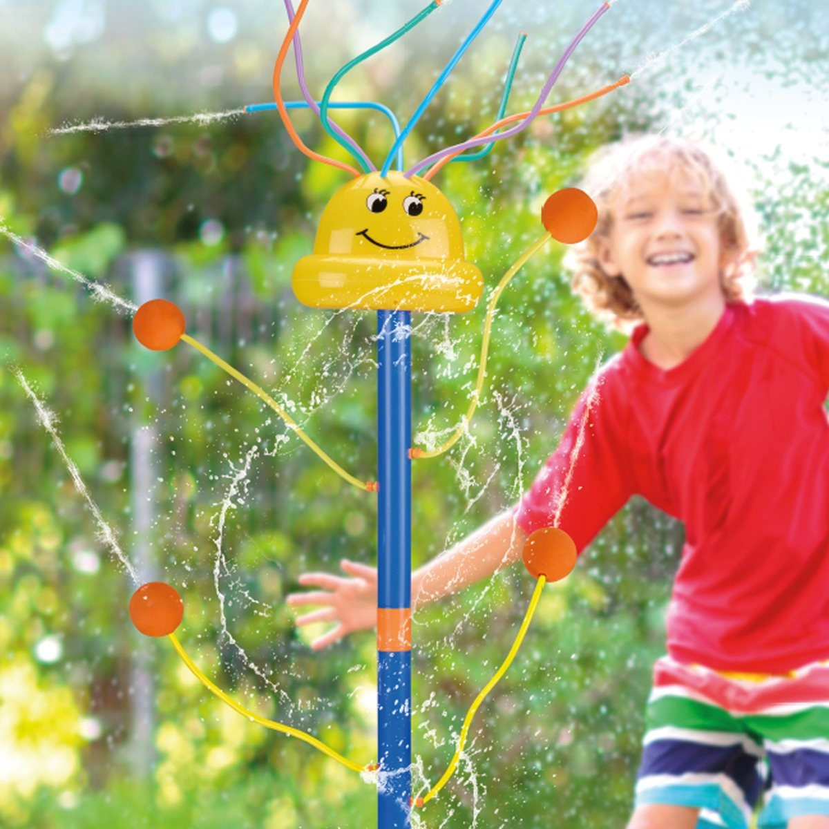 Un garçon en chemise rouge joue avec un arroseur d'eau jaune dans le jardin.