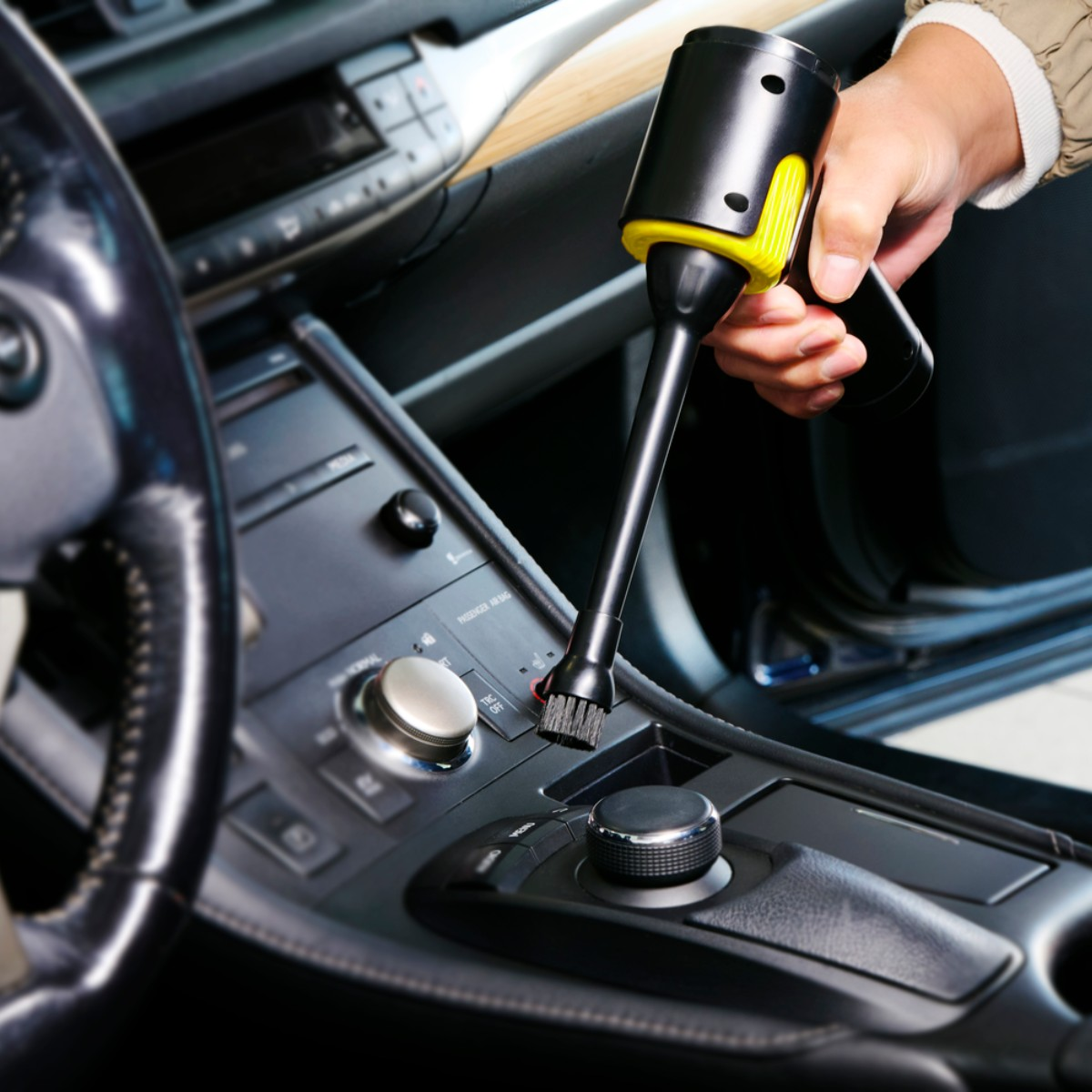 Une main utilise un petit aspirateur noir avec des accents jaunes pour nettoyer le tableau de bord d'une voiture.