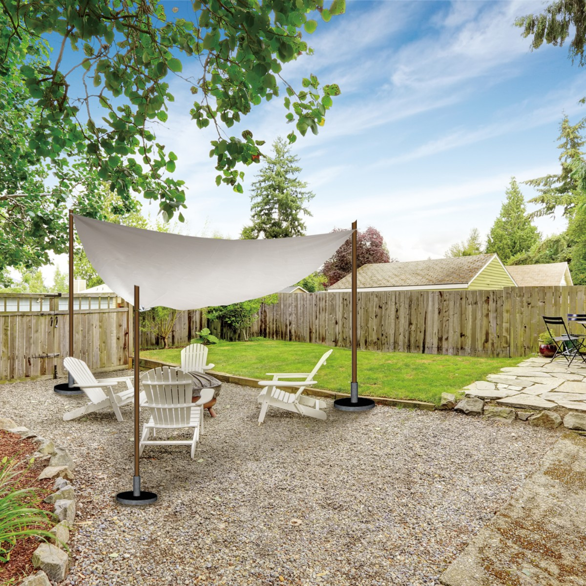 A white sunshade covers a gravel yard with four white chairs and a fire pit.