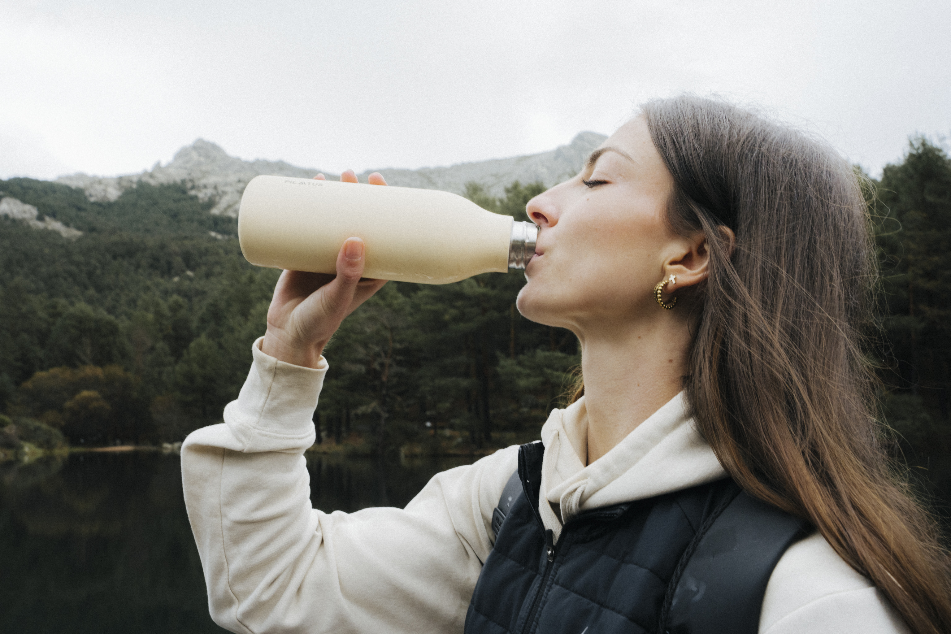 Donna beve da una bottiglia color crema davanti a un lago di montagna boscoso.