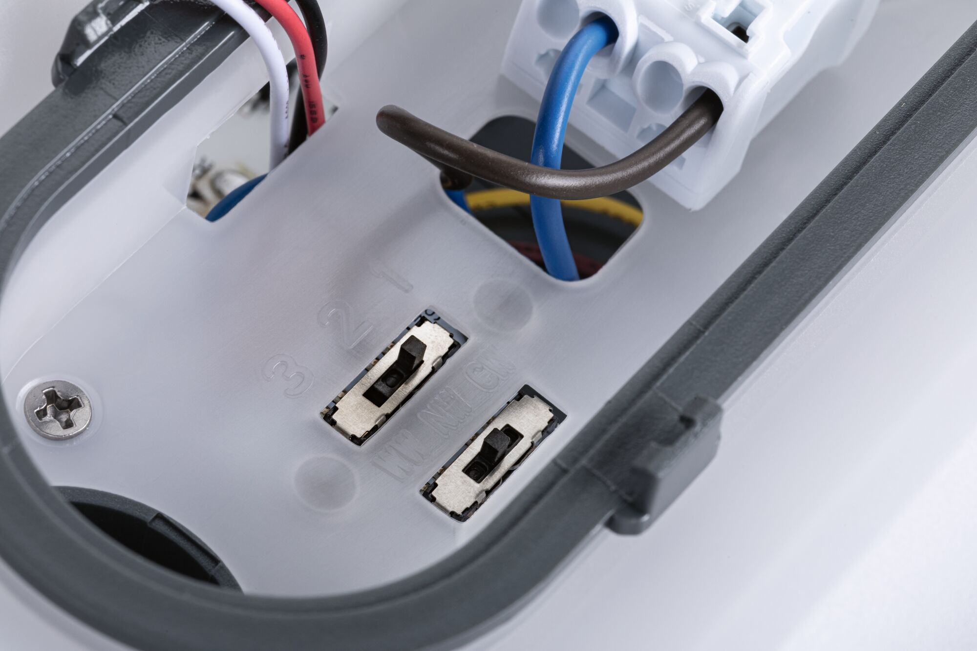 Nahaufnahme eines hellgrauen Elektrokasten-Innenraums mit verschiedenfarbigen Kabeln (rot, weiß, schwarz, blau, braun, gelb) und zwei kleinen Schaltern.