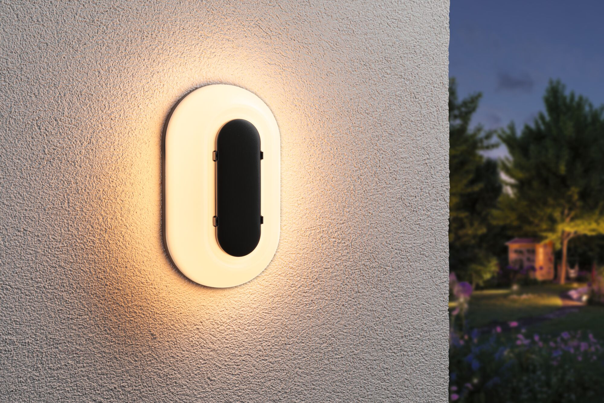 Eine ovale Wandleuchte mit schwarzem Einsatz leuchtet auf einer strukturierten Wand. Im Hintergrund ein Garten bei Nacht.
