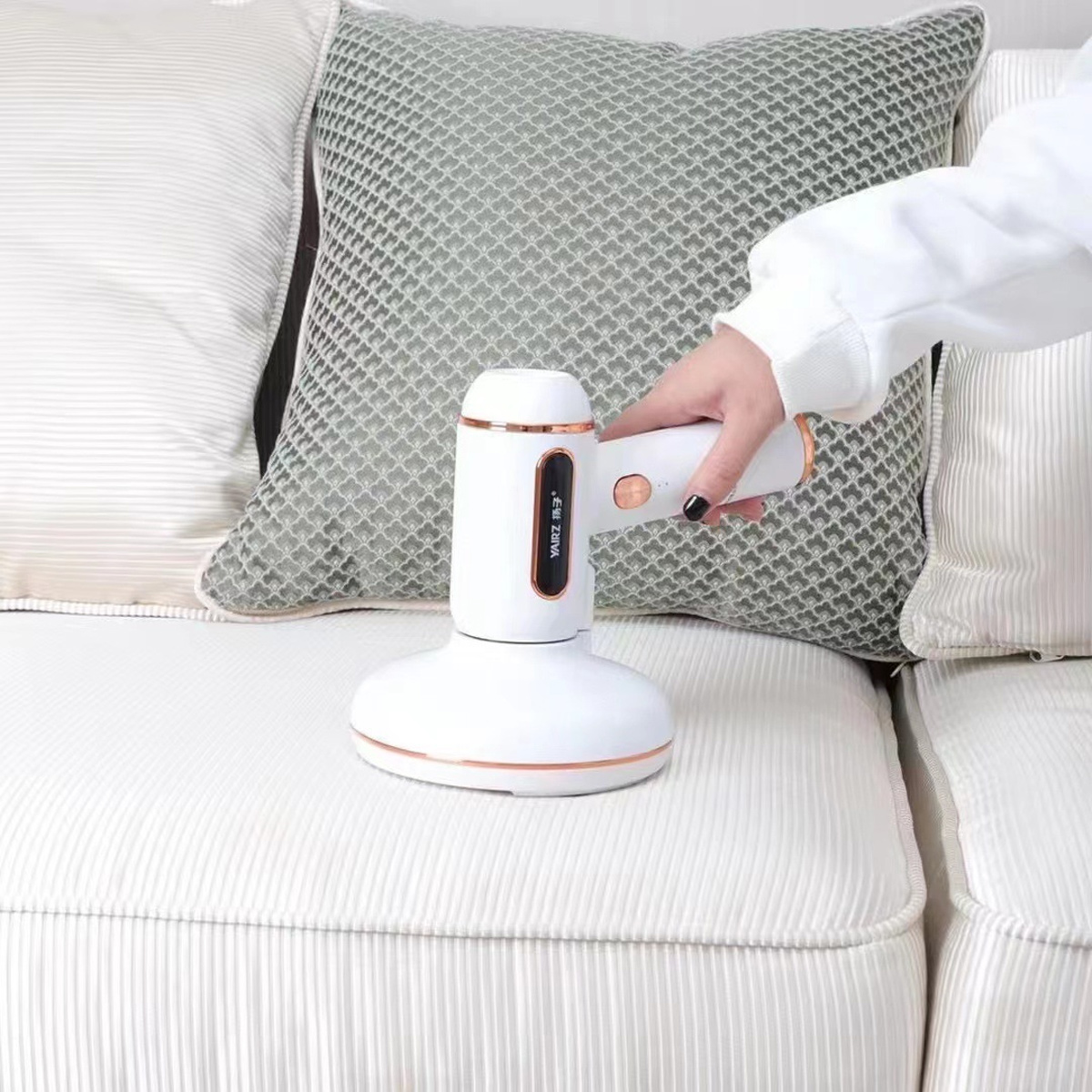 Eine weiße Hand hält einen weißen Staubsauger mit roségoldenen Akzenten auf einem weißen Sofa.