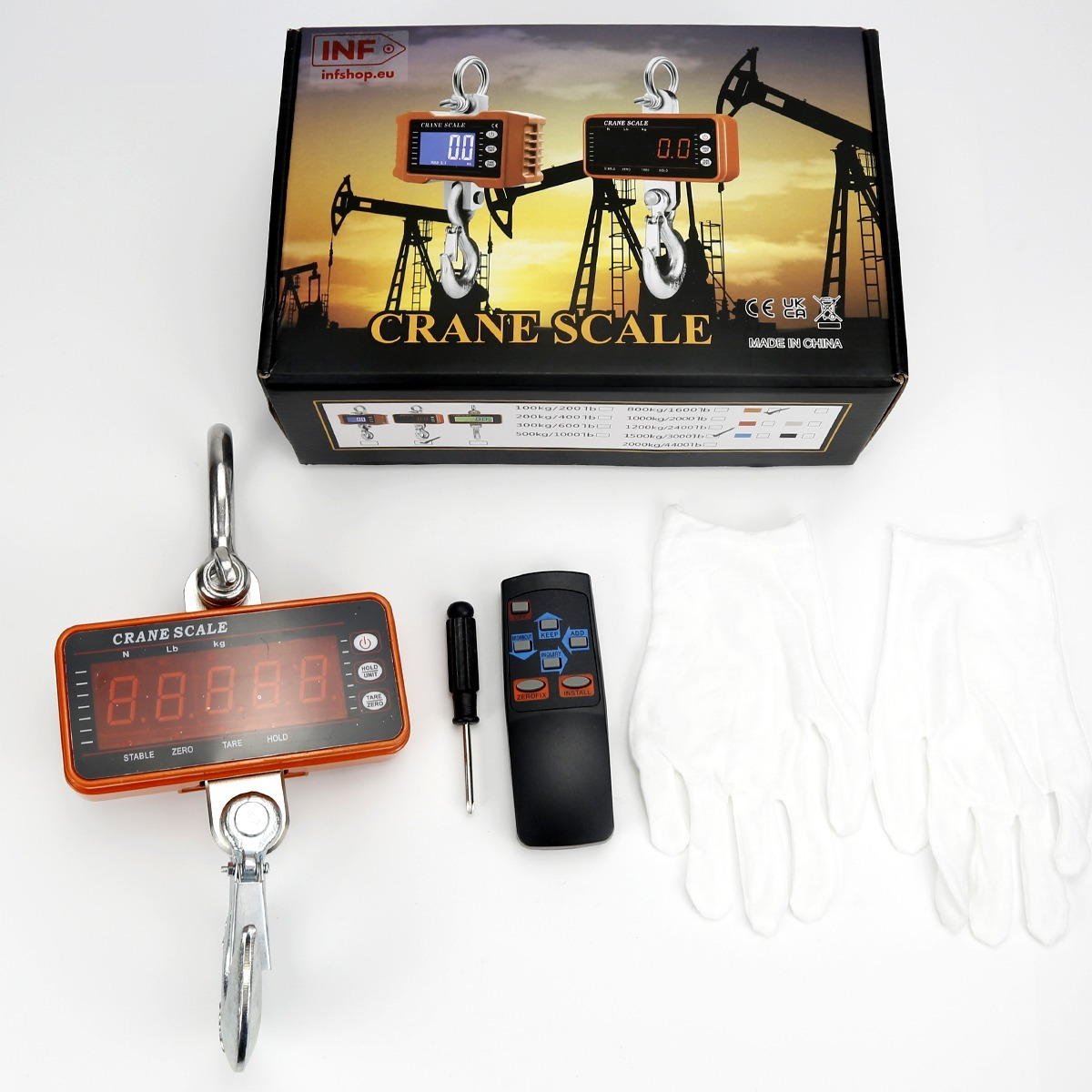 Eine Kranwaage, Fernbedienung, Schraubendreher und weiße Handschuhe liegen auf einer weißen Oberfläche, mit der Produktverpackung im Hintergrund.