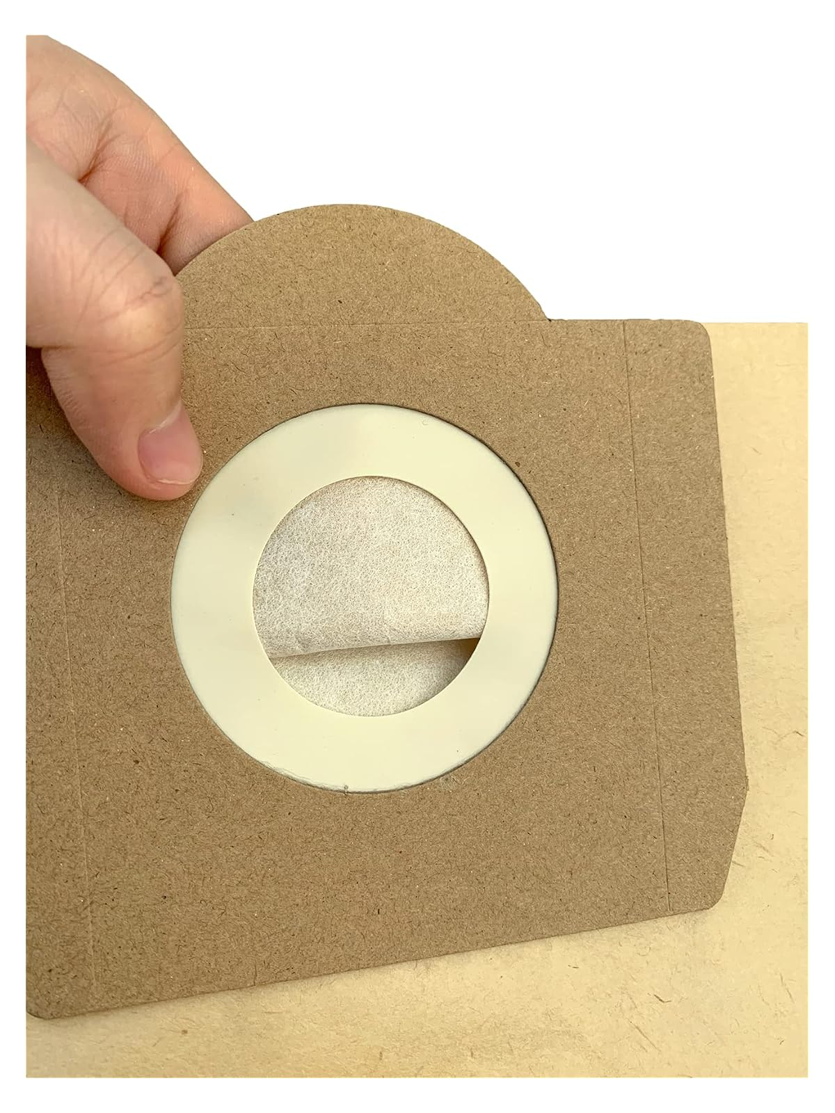 Eine Hand hält einen braunen Papier-Staubsaugerbeutel mit einer runden weißen Öffnung, die einen weißen Filter im Inneren zeigt.