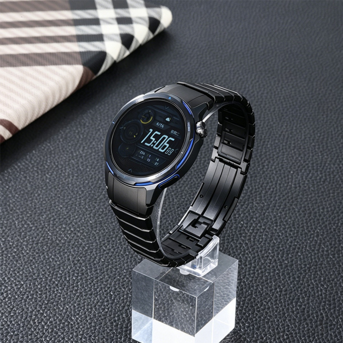 Eine schwarze Smartwatch mit blauem Akzent auf einem klaren Ständer, mit einem karierten Tuch im Hintergrund.