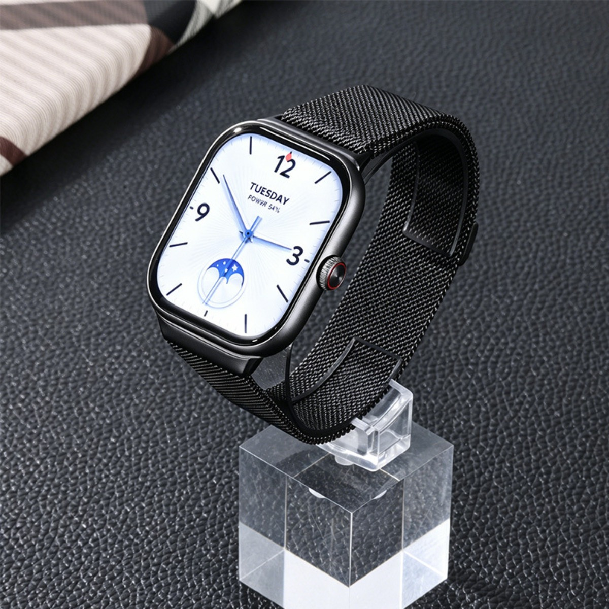 Eine schwarze Smartwatch mit Mesh-Armband wird auf einem klaren Ständer präsentiert. Der Bildschirm zeigt Uhrzeit, Datum, Akku.