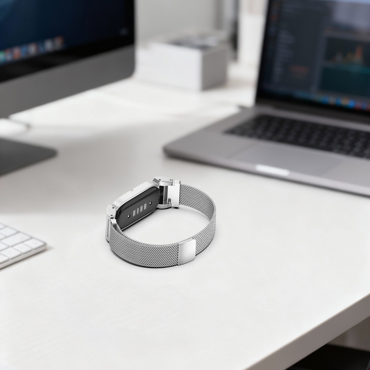Eine silberne Smartwatch mit Mesh-Armband liegt auf einem weißen Schreibtisch neben einem Laptop.