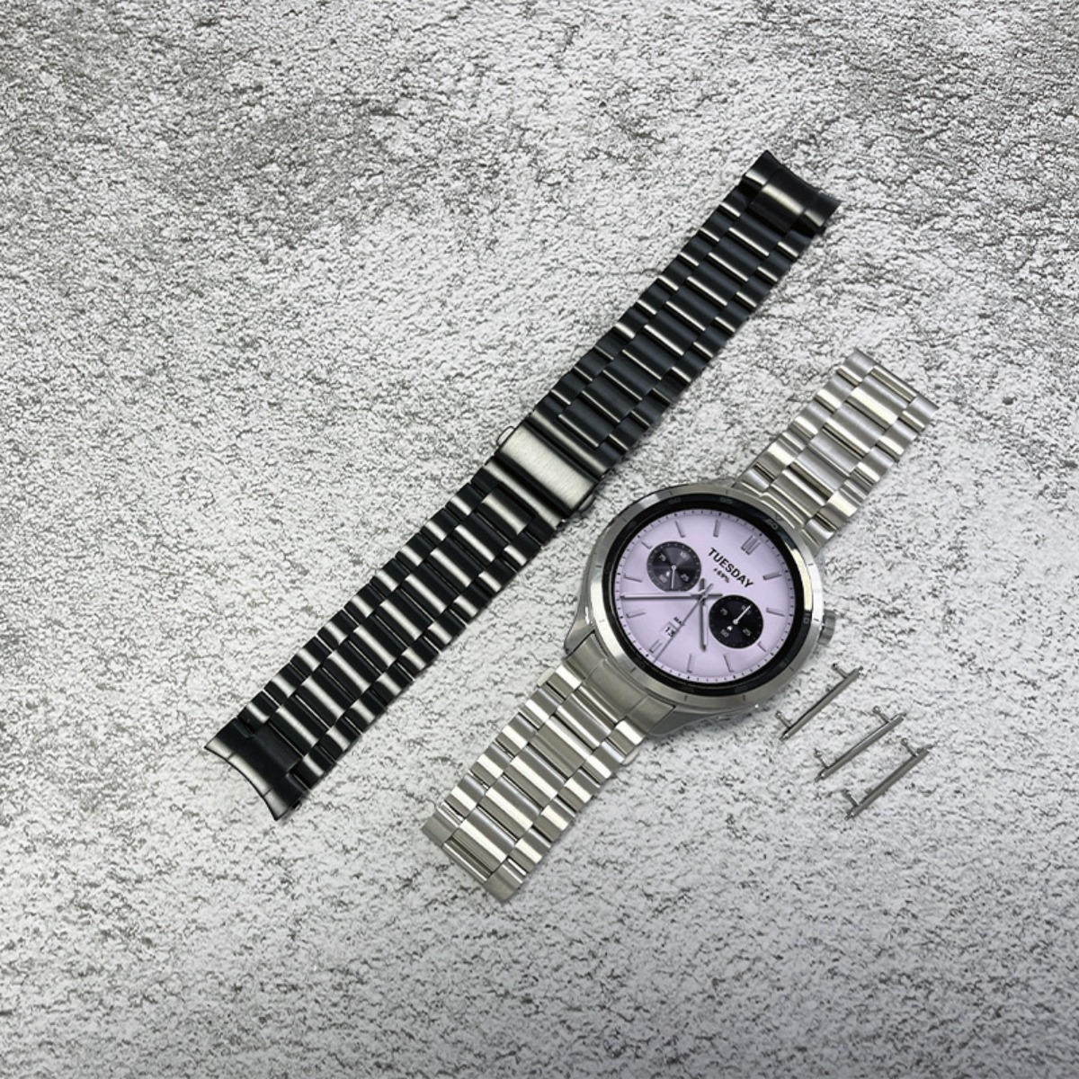 Silberne Smartwatch mit hellvioletter Zifferblatt und schwarzem Uhrenarmband daneben.