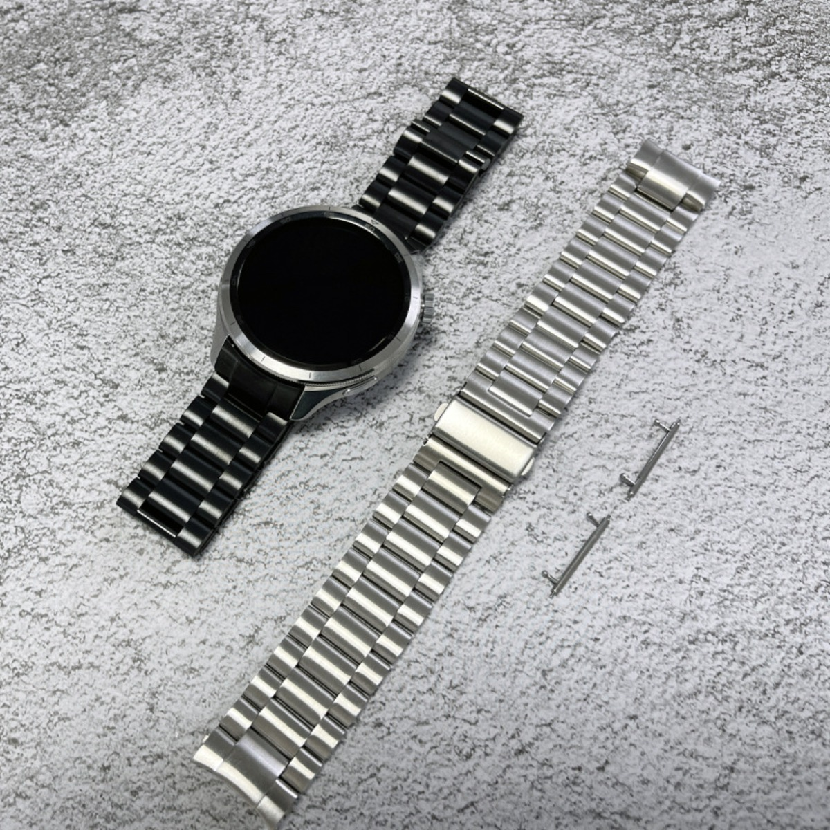 Eine schwarze und eine silberne Uhrenarmband liegen auf grauem Untergrund.