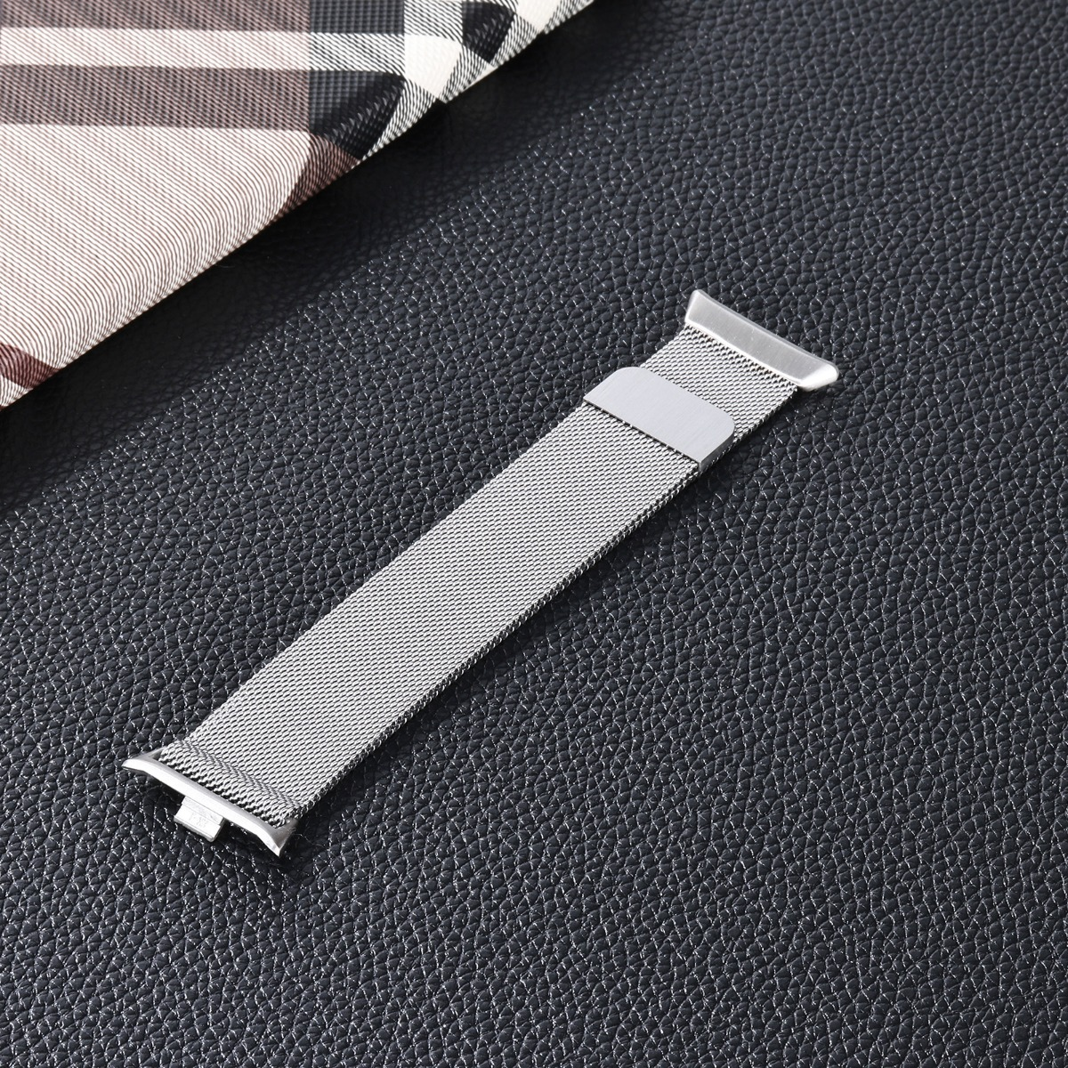 Ein silbernes Milanaise-Uhrenarmband mit Schließe liegt diagonal auf einer schwarzen strukturierten Oberfläche, mit einem karierten Stoff oben links.