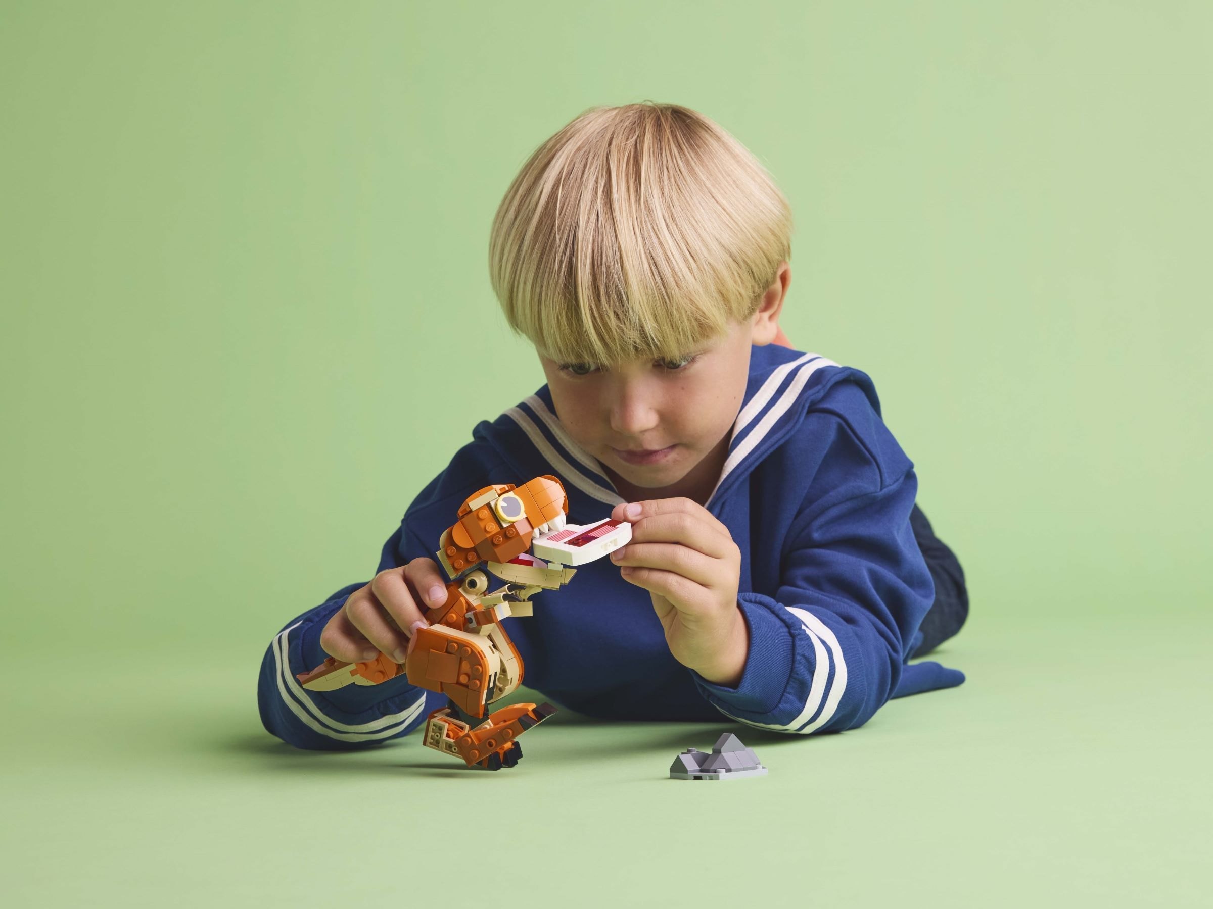 Ein kleiner Junge mit blonden Haaren in einem blauen Oberteil liegt auf einer hellgrünen Fläche und spielt mit einem orangefarbenen und beigen LEGO-Dinosaurier.