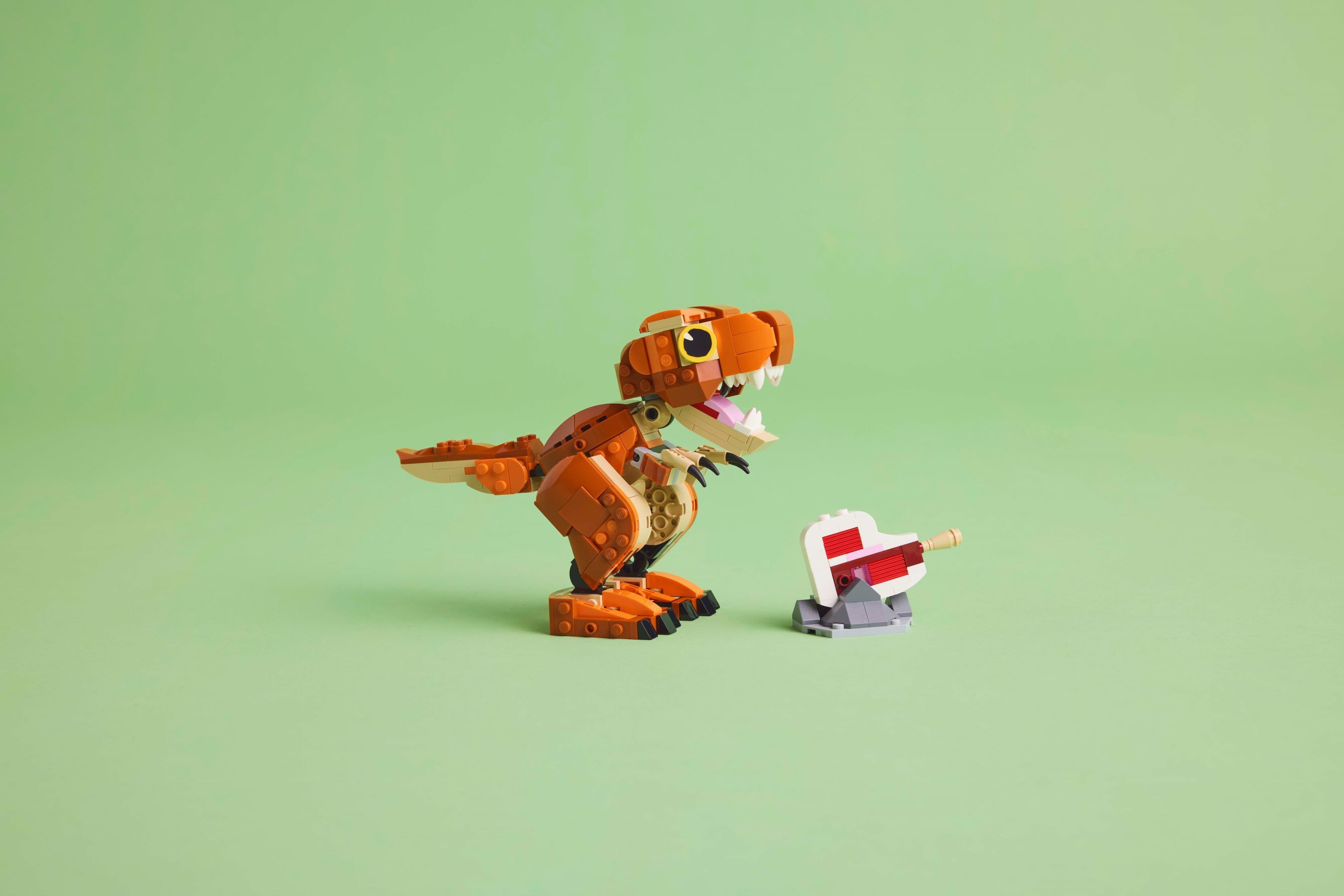 Ein orangefarbener Lego-Dinosaurier steht neben einer weißen und roten Lego-Kanone auf einem hellgrünen Hintergrund.