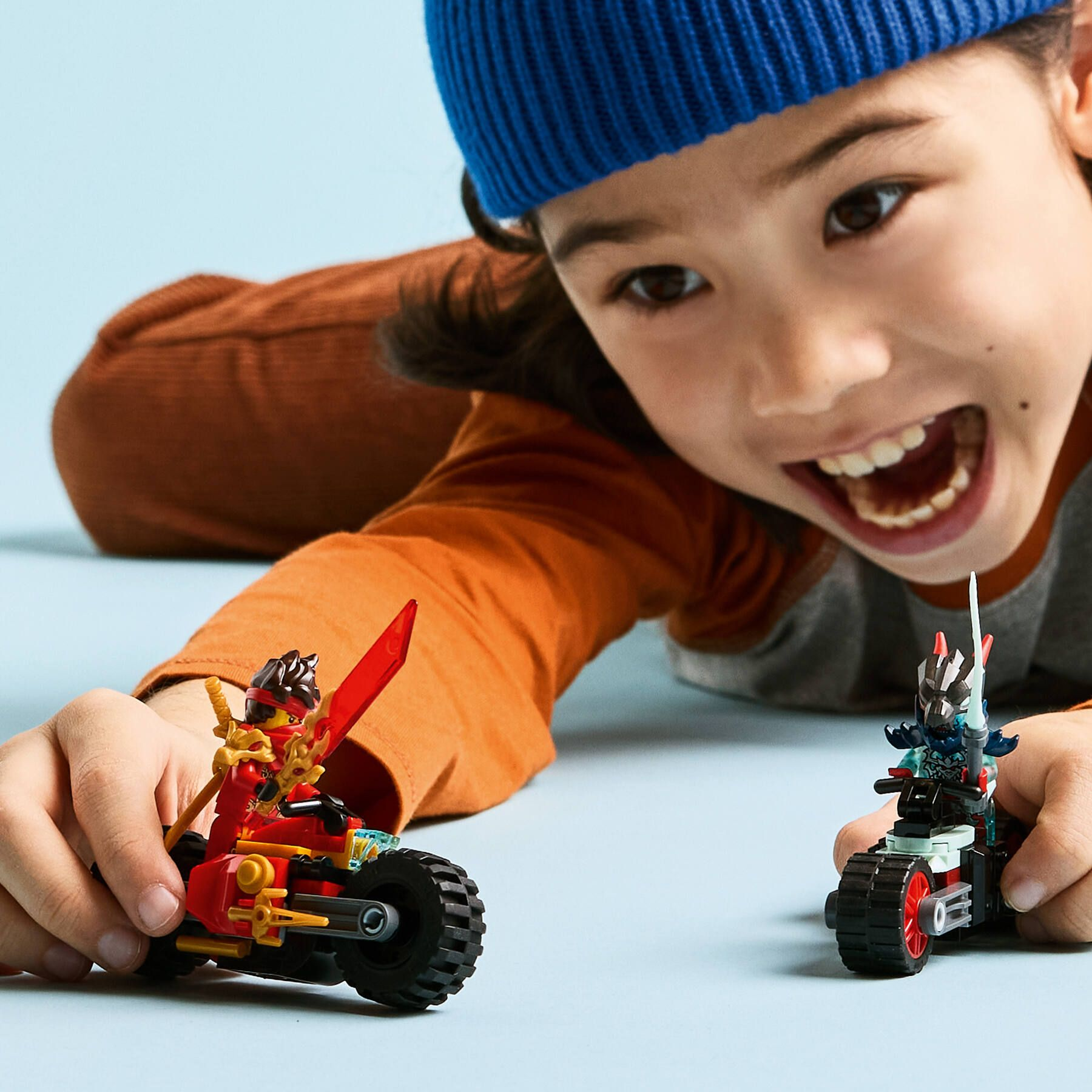 Bambino con moto Lego Ninjago: una rossa, una blu, su sfondo azzurro.