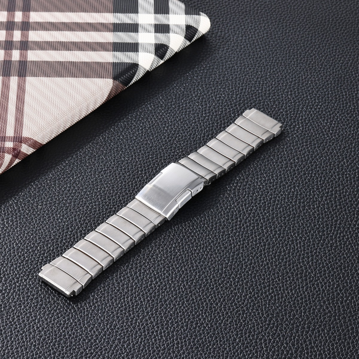 Ein silbernes Metalluhrenarmband liegt auf schwarzem Leder neben kariertem Stoff.