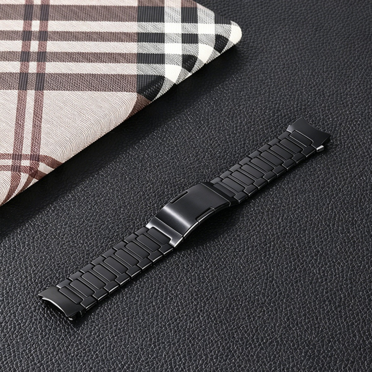 Ein schwarzes Uhrenarmband aus Metall liegt auf schwarzem Leder neben einem karierten Stoff.
