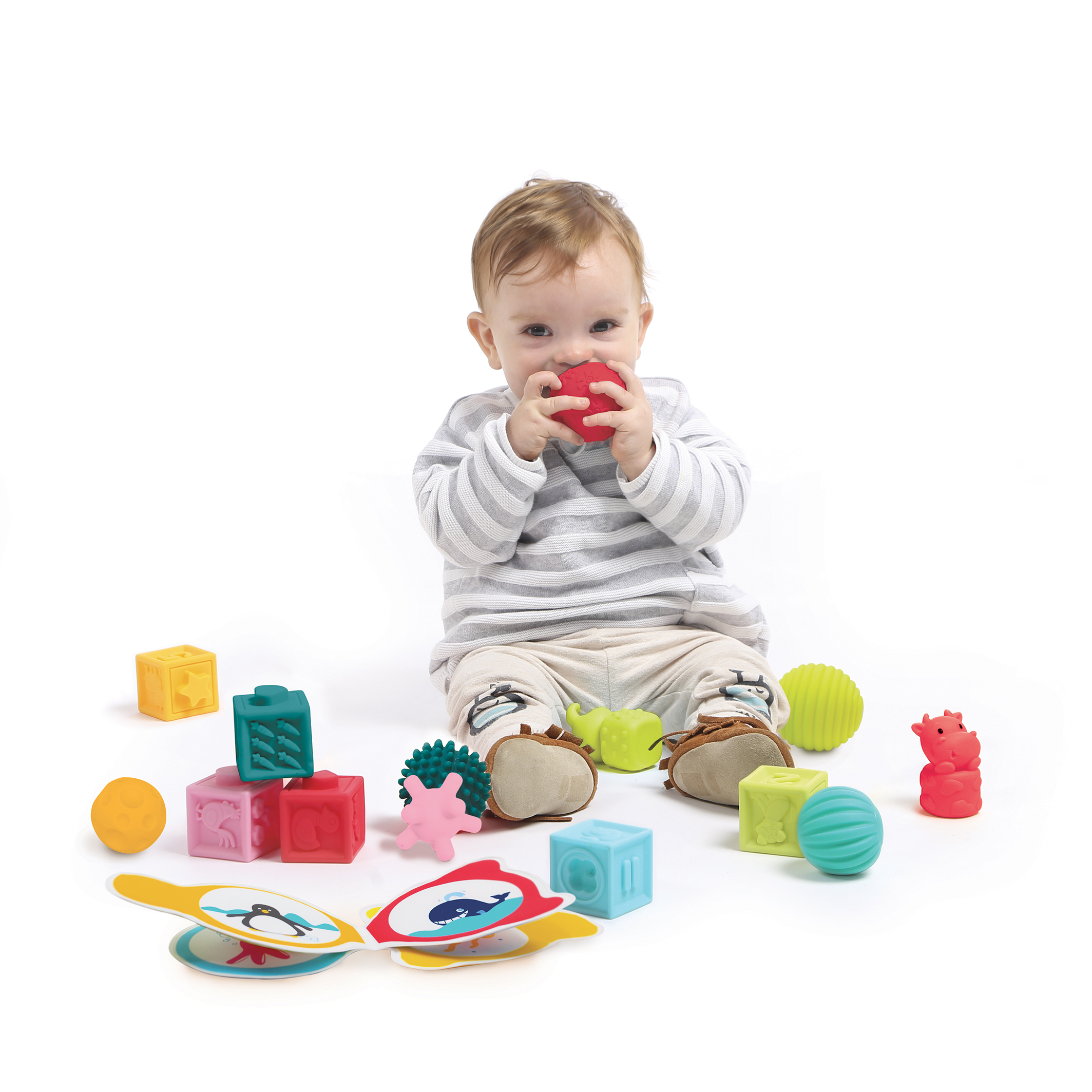 Ein Baby sitzt zwischen bunten Babyspielzeugen auf weißem Hintergrund.