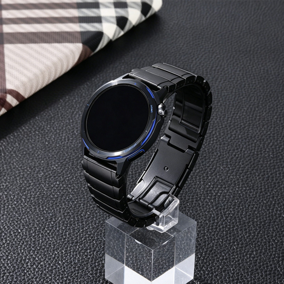 Schwarze Smartwatch mit Metallband auf klarem Ständer, neben gemustertem Tuch.