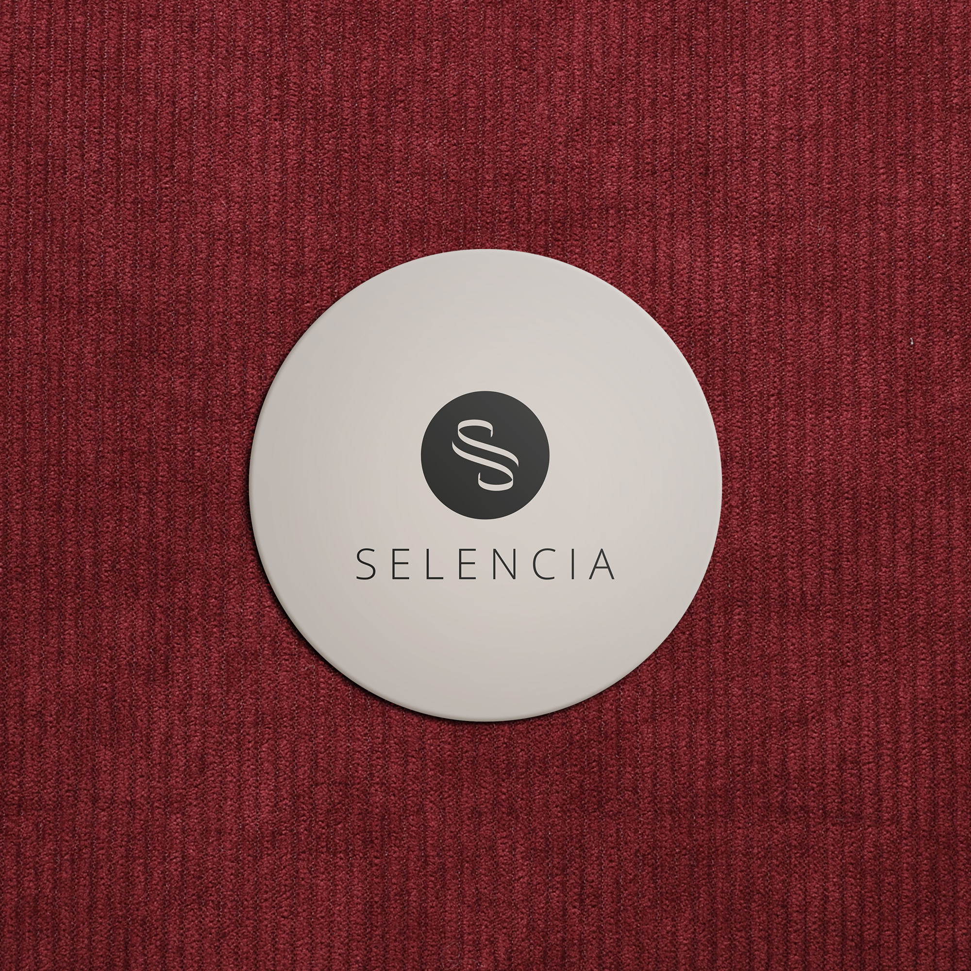 Ein weißer runder Untersetzer mit schwarzem 'S'-Logo und Schriftzug SELENCIA auf rotem Stoff.