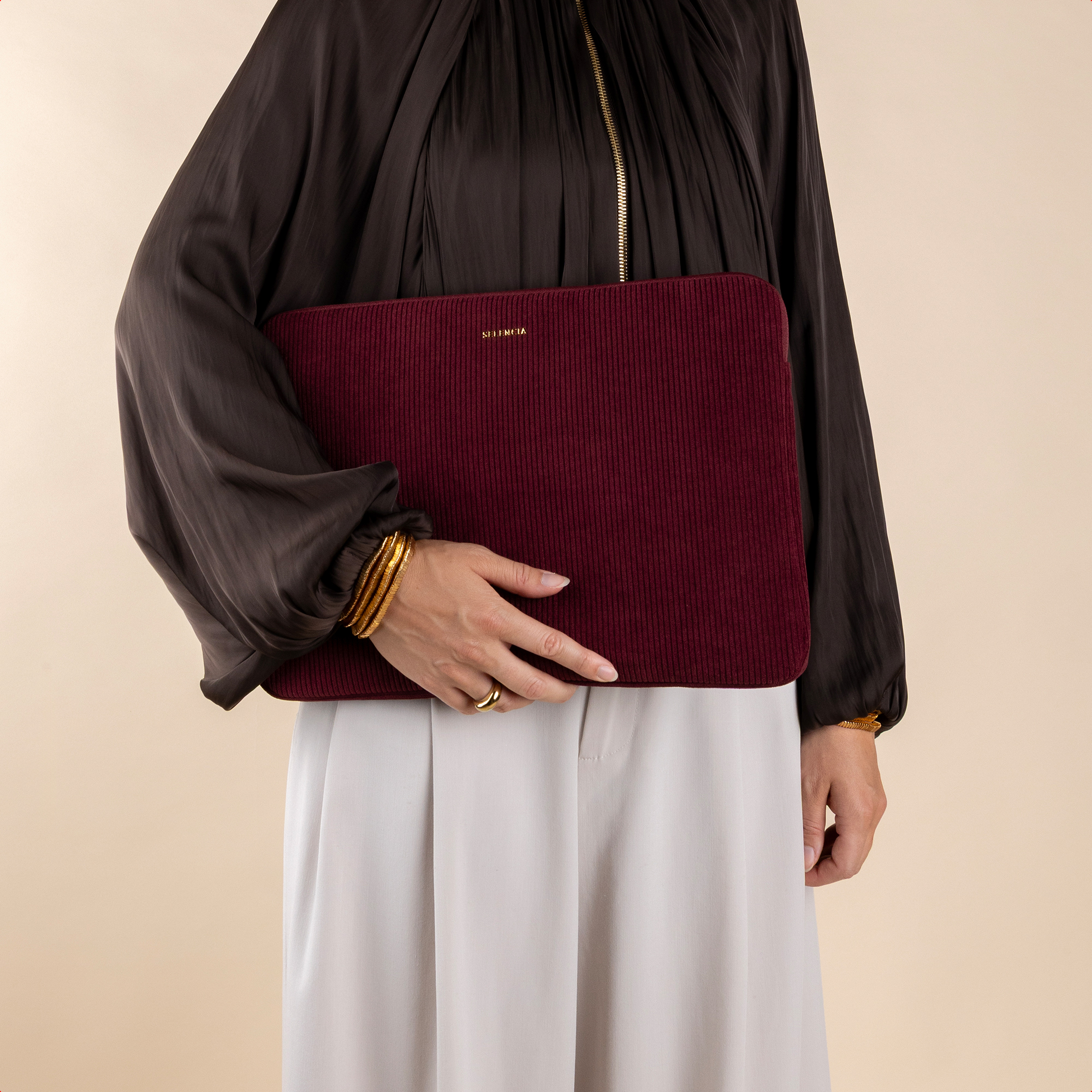 Eine Person hält eine burgunderrote Laptoptasche mit Rillen. Sie trägt eine dunkle Bluse und helle Hosen.